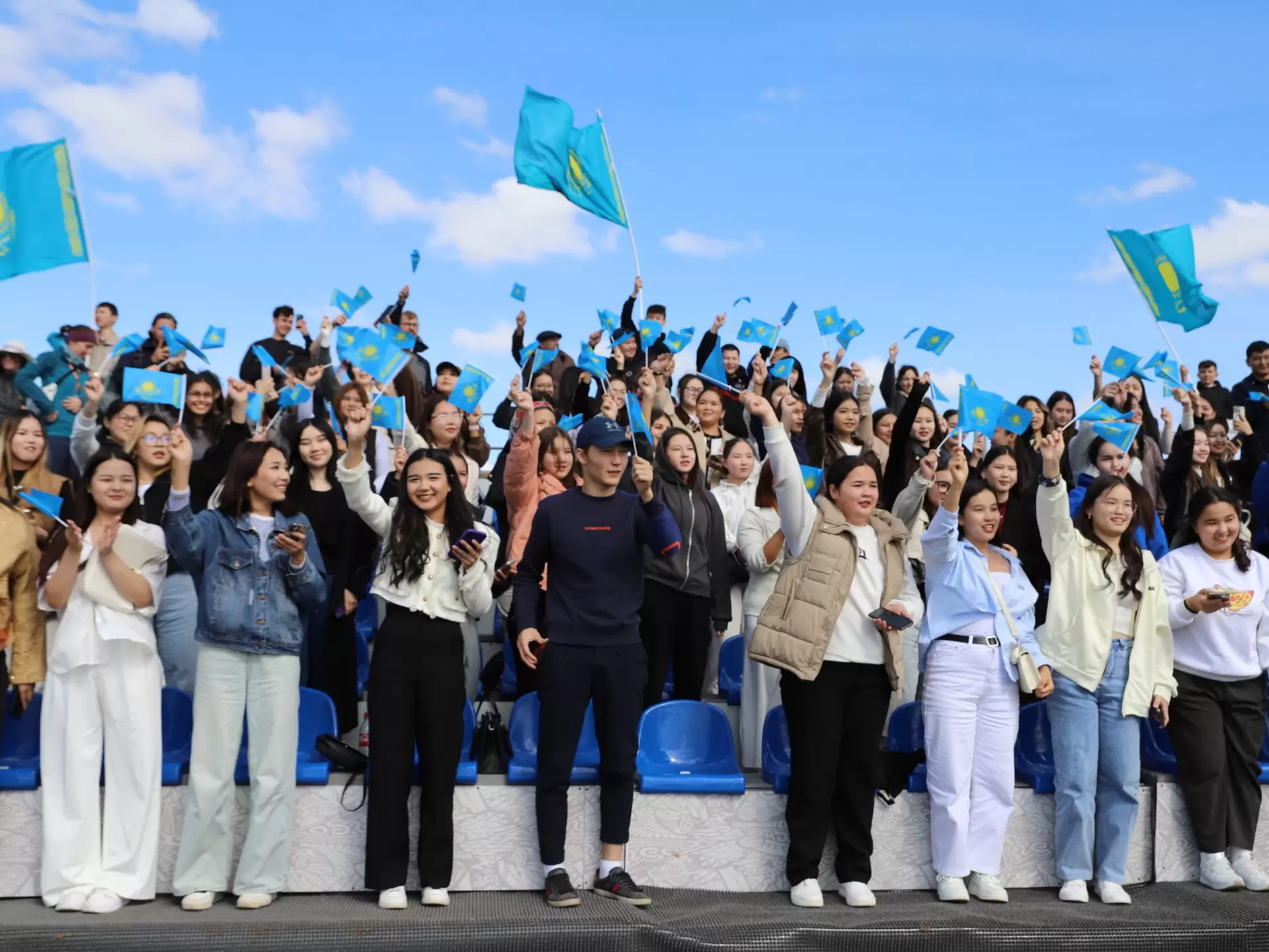 Groups of spectators cheer and wave Kazakhstan's blue and yellow national flag on the side of the game arena at the World Nomad Games in 2024 