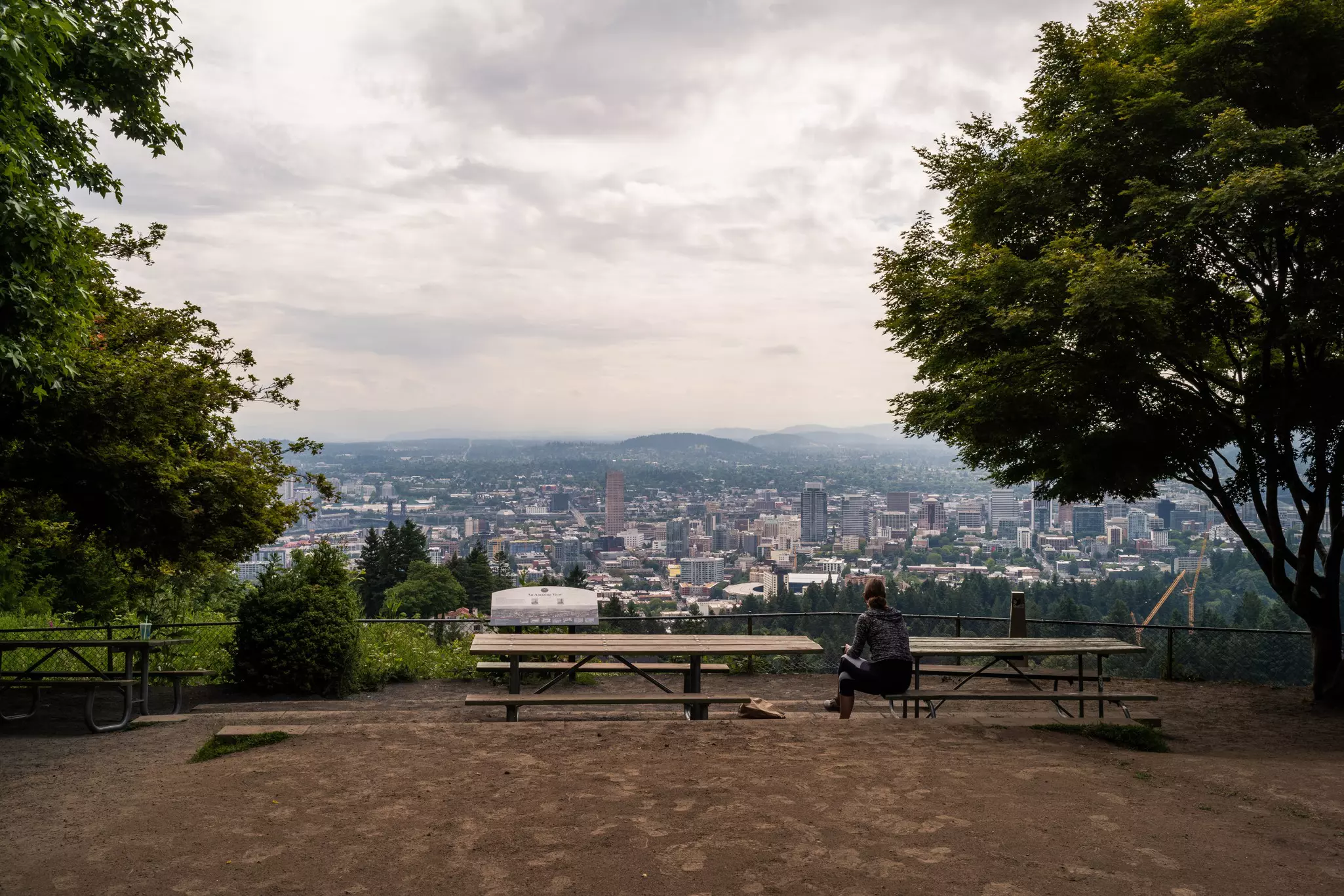 A person sits at a picnic table overlooking a city skyline on a cloudy day.