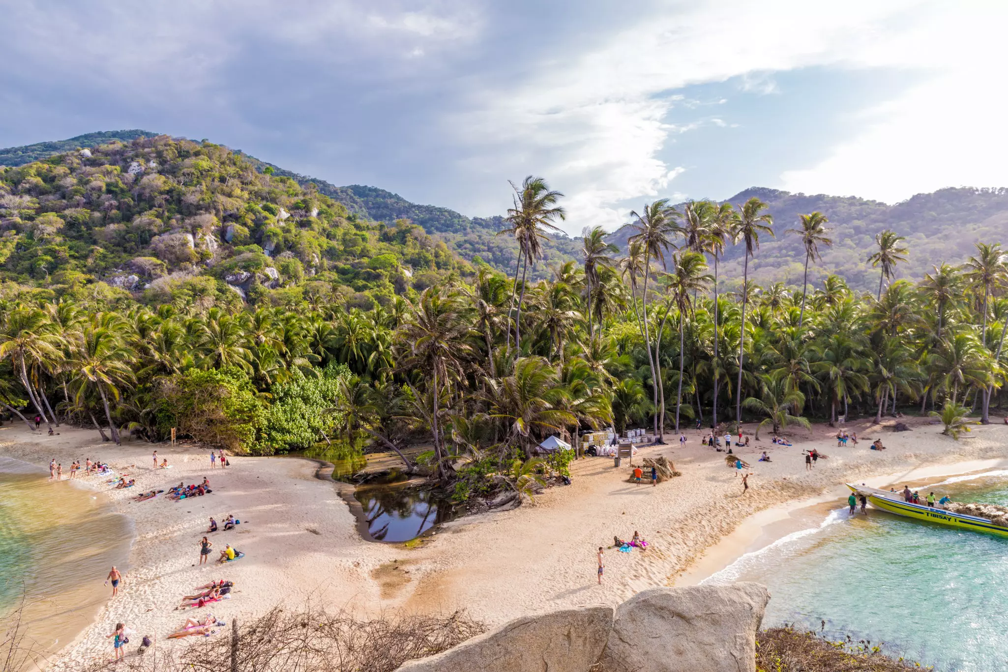People relax on a sandy tropical beach backed by dense jungle and tall palm trees.