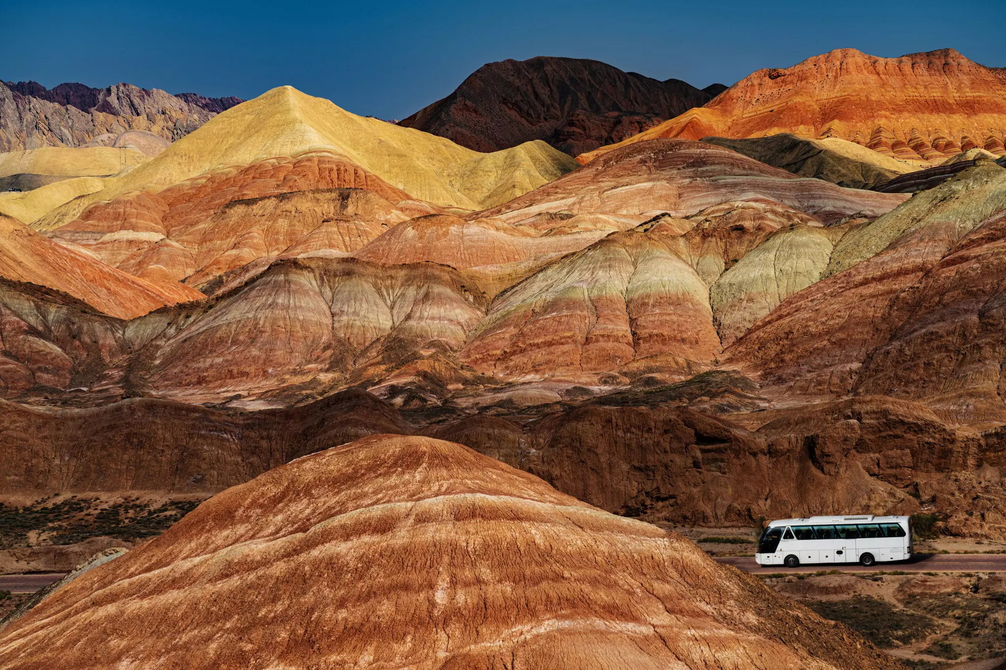 A white bus drives by mountains with rocks in hues of gold, orange and red.