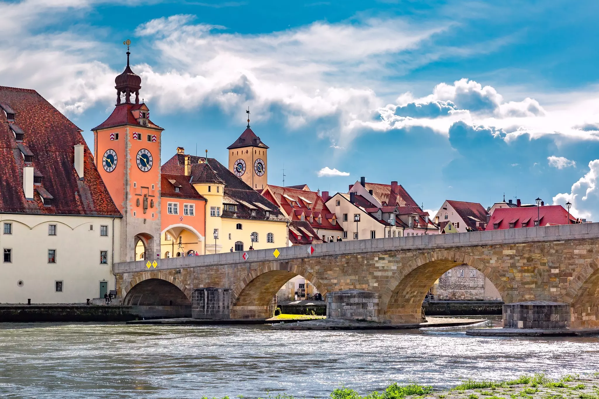 A stone bridge with several piers spans a river. Historic buildings are seen along the riverfront.