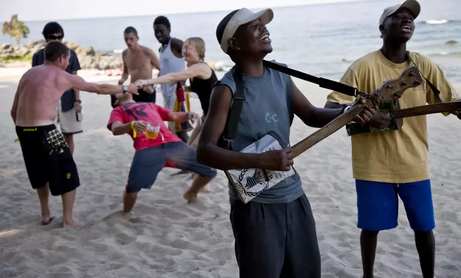 Malawian men singing and strumming home-made tin guitars on a Lake Malawi beach as travellers dance behind