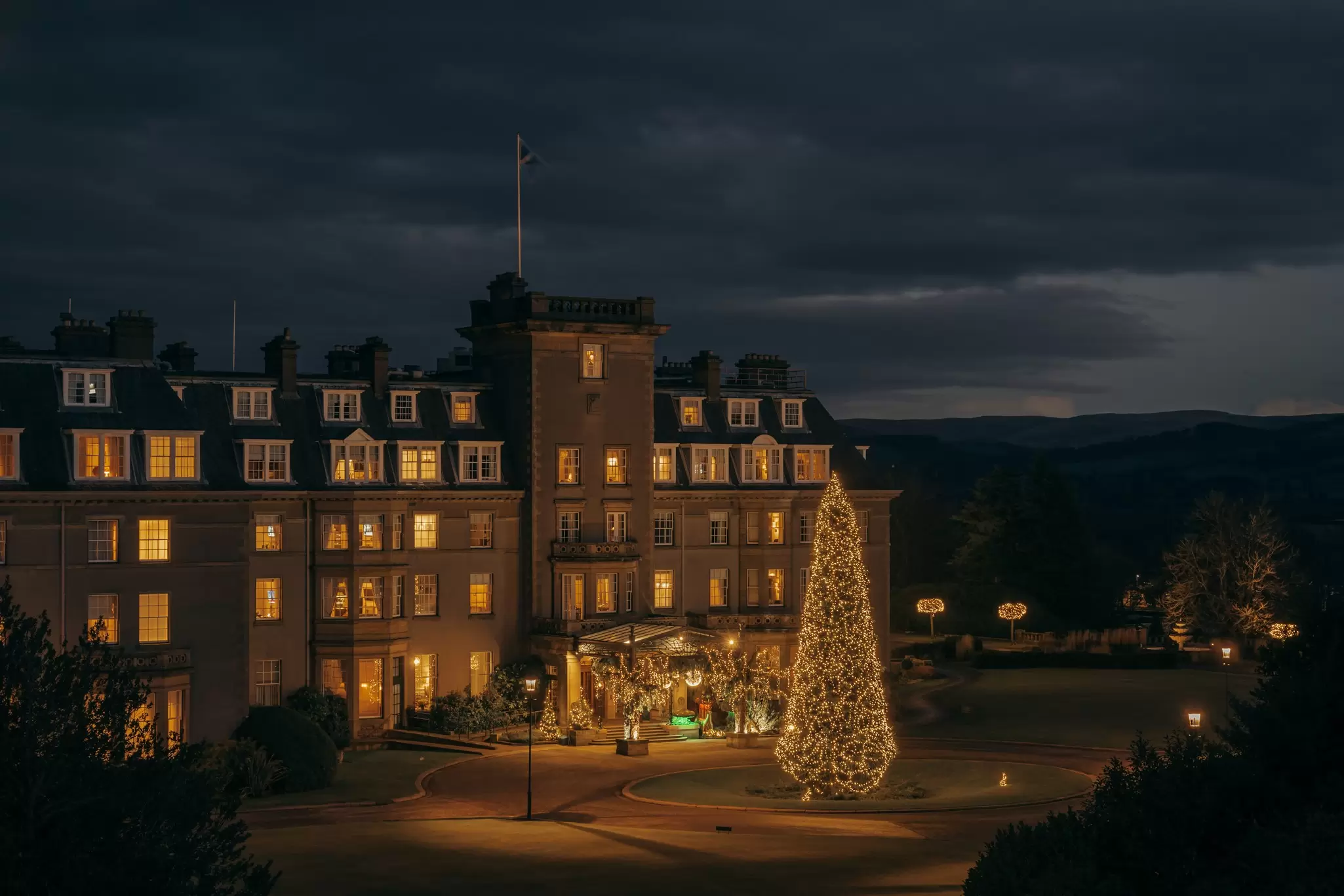 Exterior of a rural a hotel at night, with a giant lit-up Christmas tree in the front yard and twinkling garland framing the front entrance