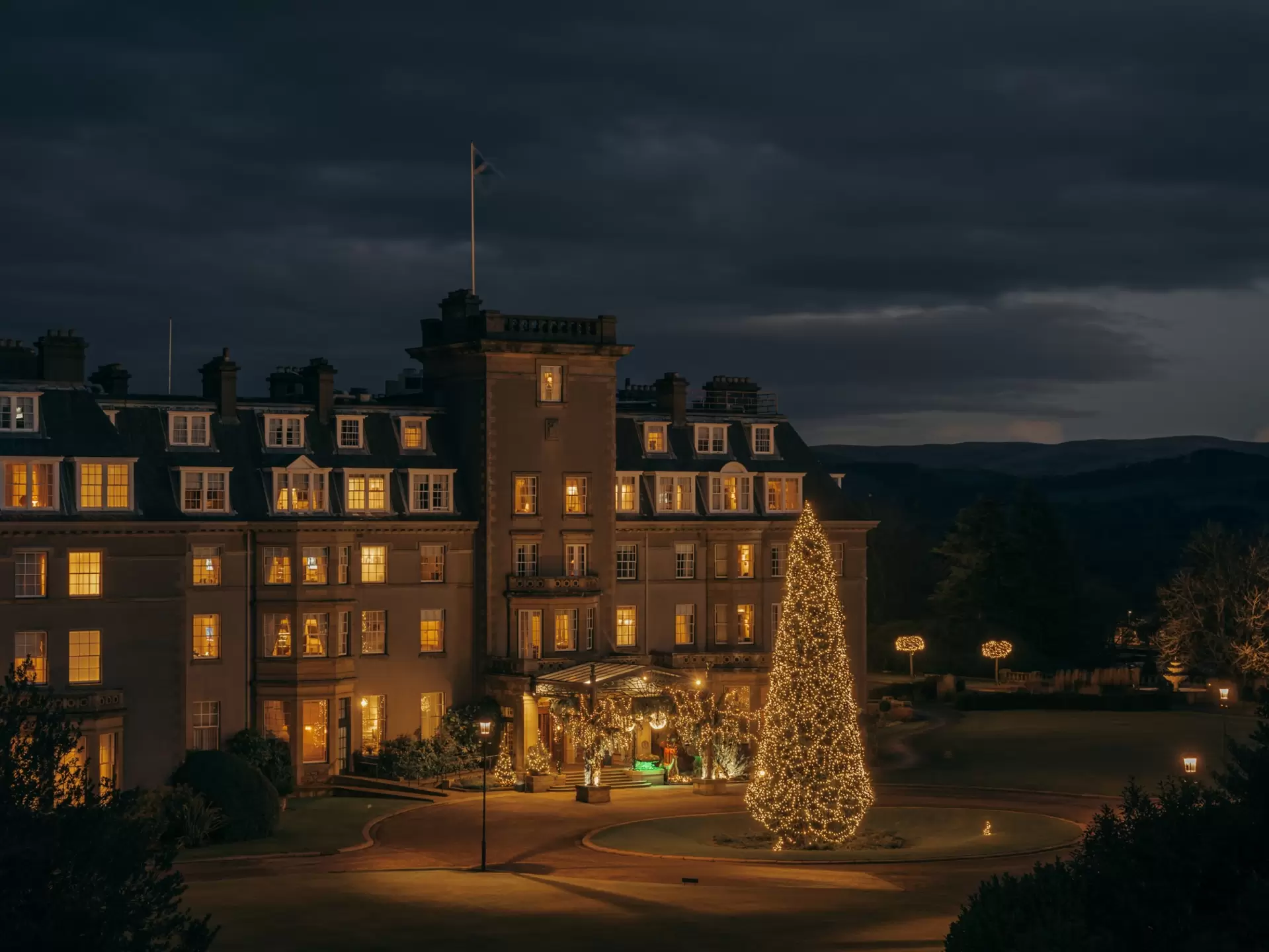 Exterior of a rural a hotel at night, with a giant lit-up Christmas tree in the front yard and twinkling garland framing the front entrance