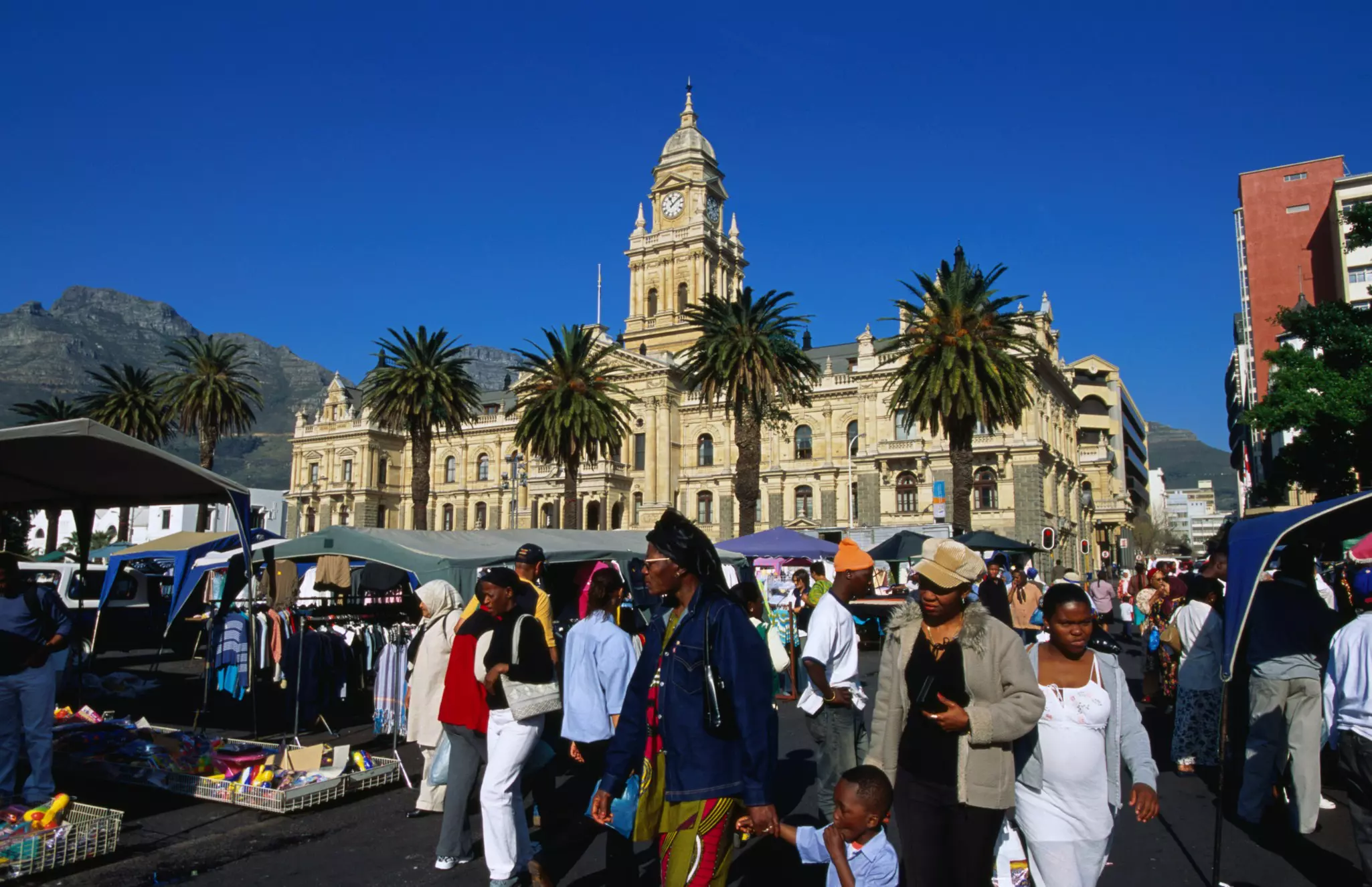 A crowd of people walks by a vendors in a market in a busy city square. An ornate building and palm trees are seen on the edge of the square.