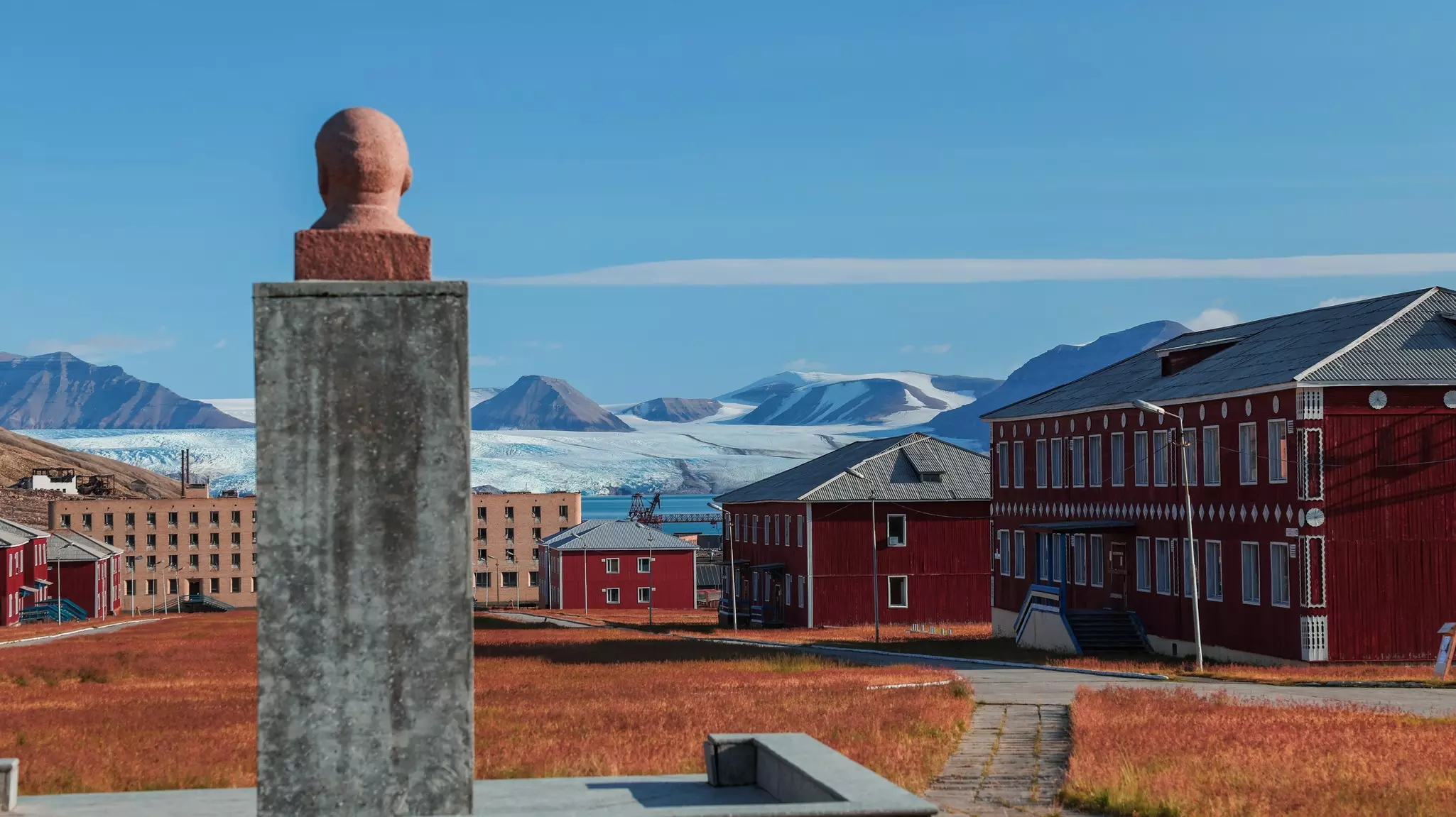 A bust of a human head on a plinth is seen from beyond, with the buildings of an abandoned settlement pictured at the other side of a brown lawn. Frozen landscapes with mountains are seen in the distance.