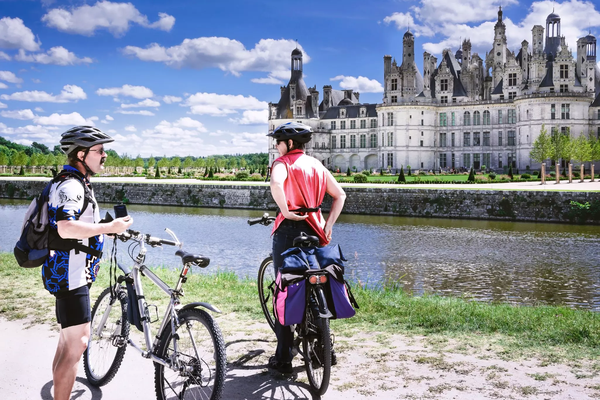 Two cyclists in athletic gear rest by a bike path on a river. An ornate château stands across the river.