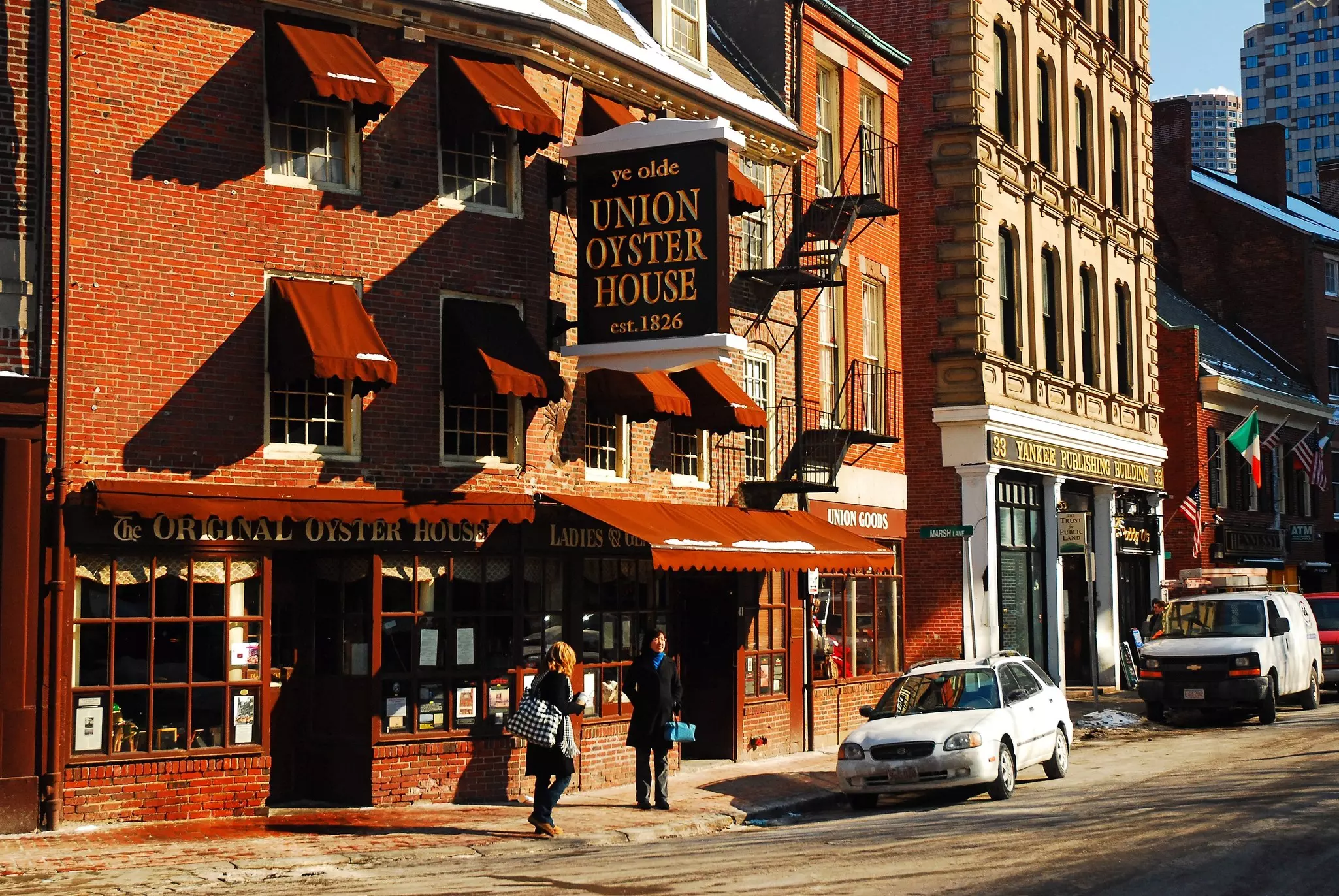Two people walk by a red-brick restaurant with a large sign that says "Ye Olde Union Oyster House, est. 1826" on a sunny winter's day.