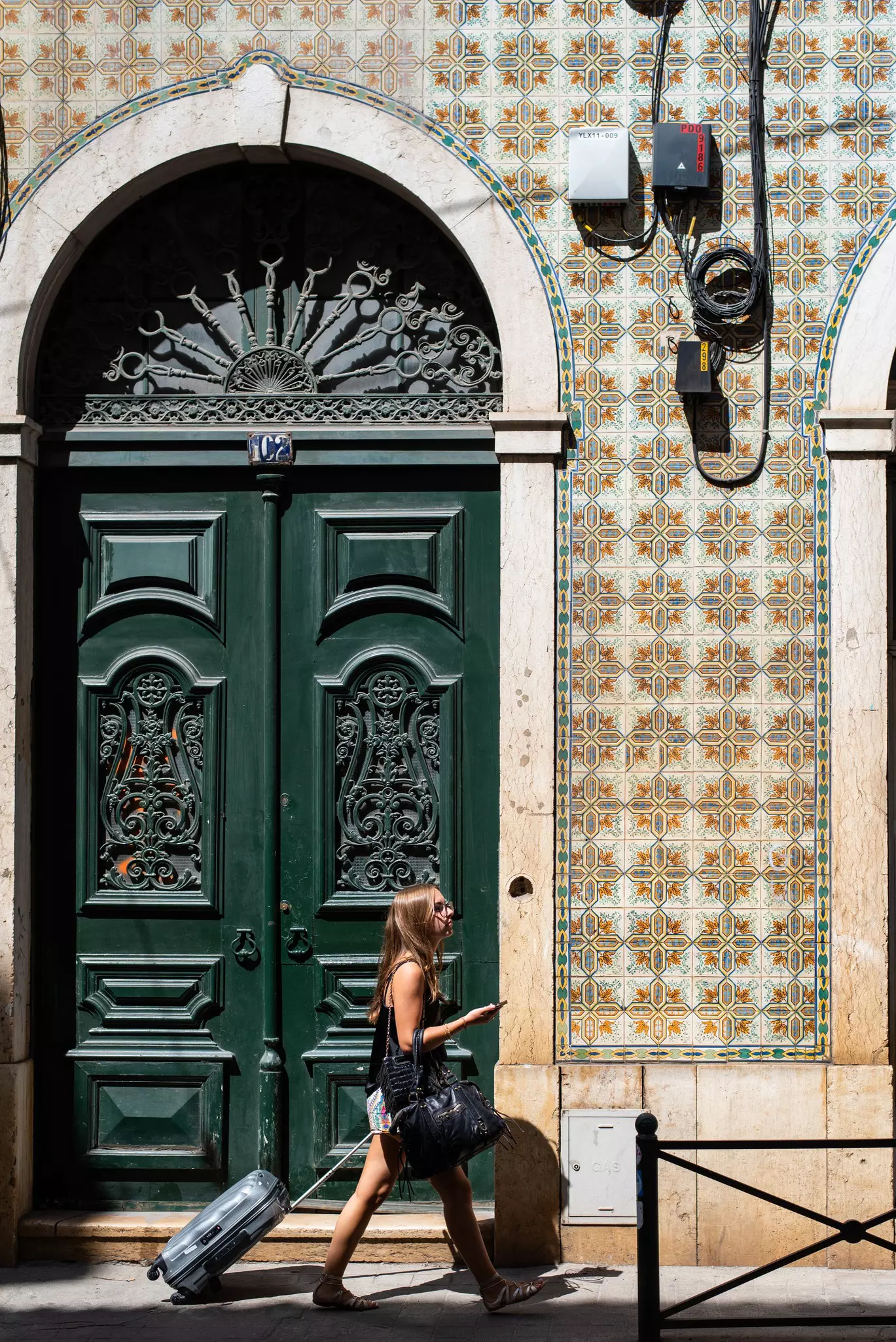 A woman walks with a rolling suitcase in front of a tiled wall in Santos, Lisbon