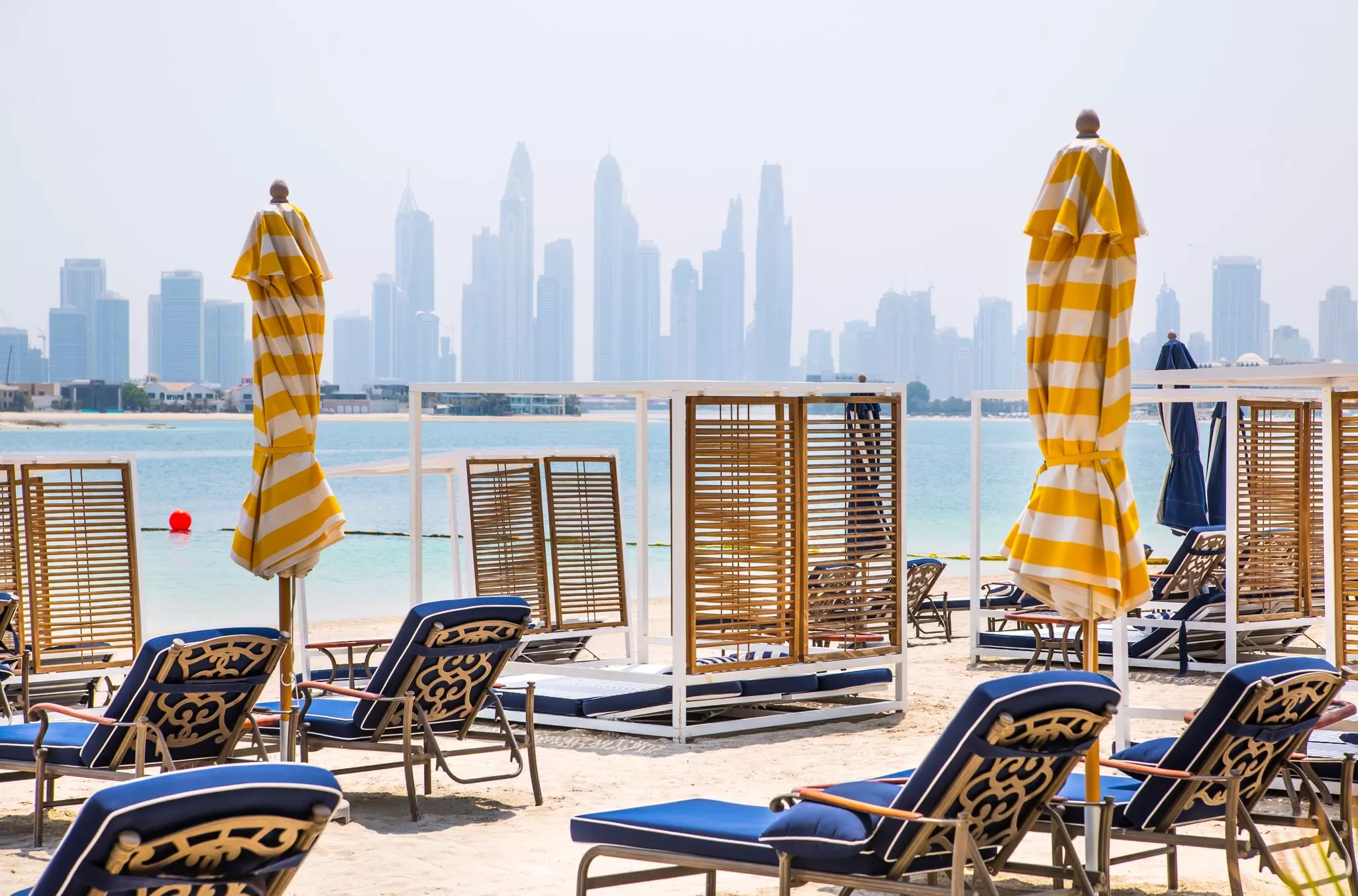 Blue lounge chairs are set up on white sand, with closed yellow-and-white striped umbrellas between them; the skyline of Dubai, United Arab Emirates, is visible across a pale blue stretch of water.