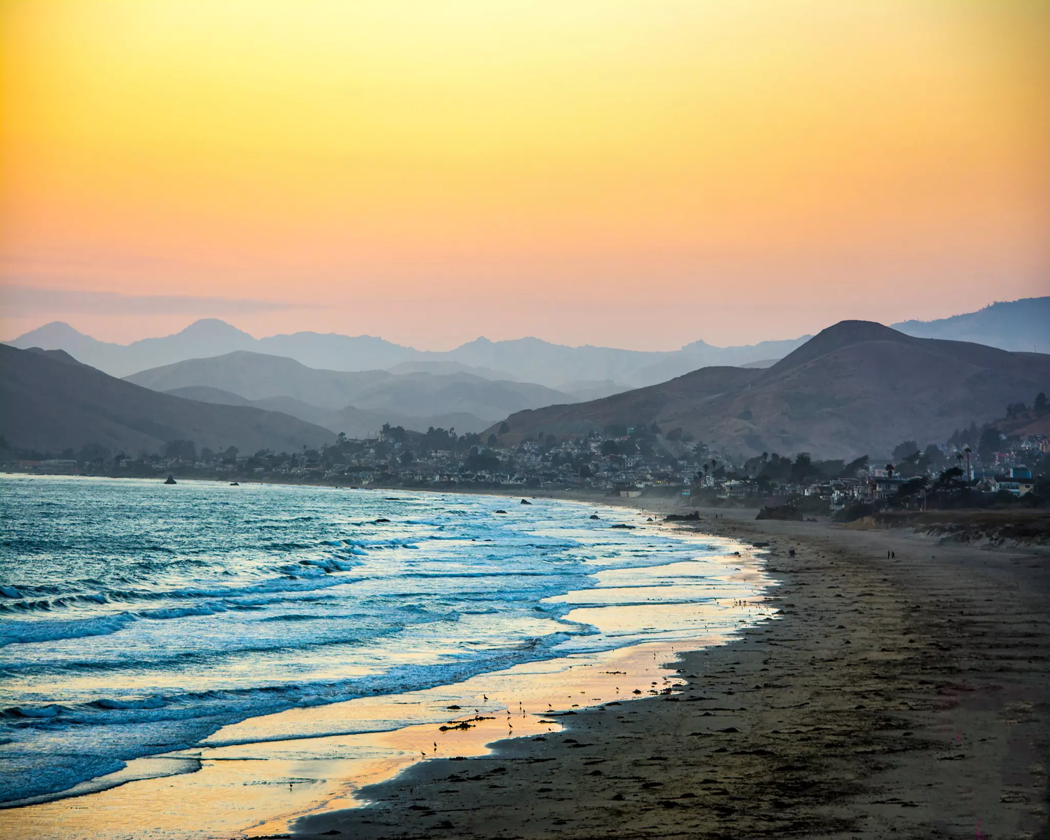Waves lap a beach just after sunset. The water appears bright blue, and shades of pink, orange and purple are visible set against mountains in the distance.