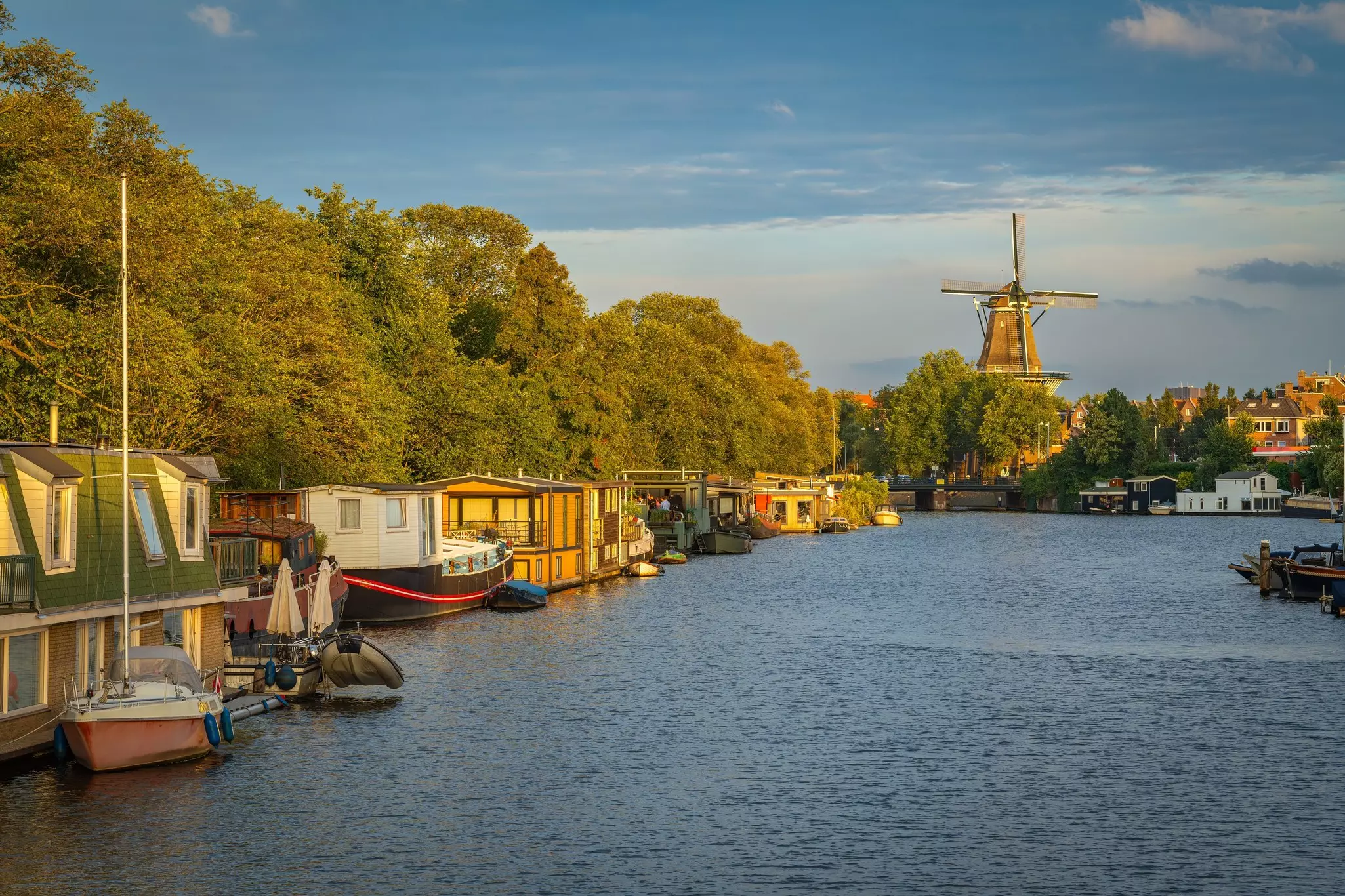 A windmill towers over leafy trees and low-rise buildings at the end of a waterway with houseboats.