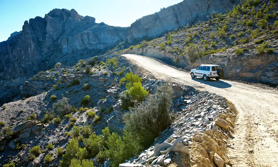 White 4WD on a gravel track below limestone cliffs on Jebel Shams, Oman, with green scrub on rocky slopes
