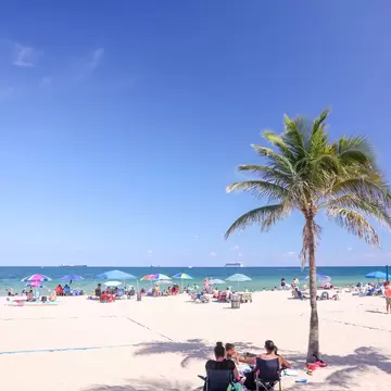 Two people in beach chairs in palm shade on white sand, colourful umbrellas and turquoise Atlantic beyond, Fort Lauderdale