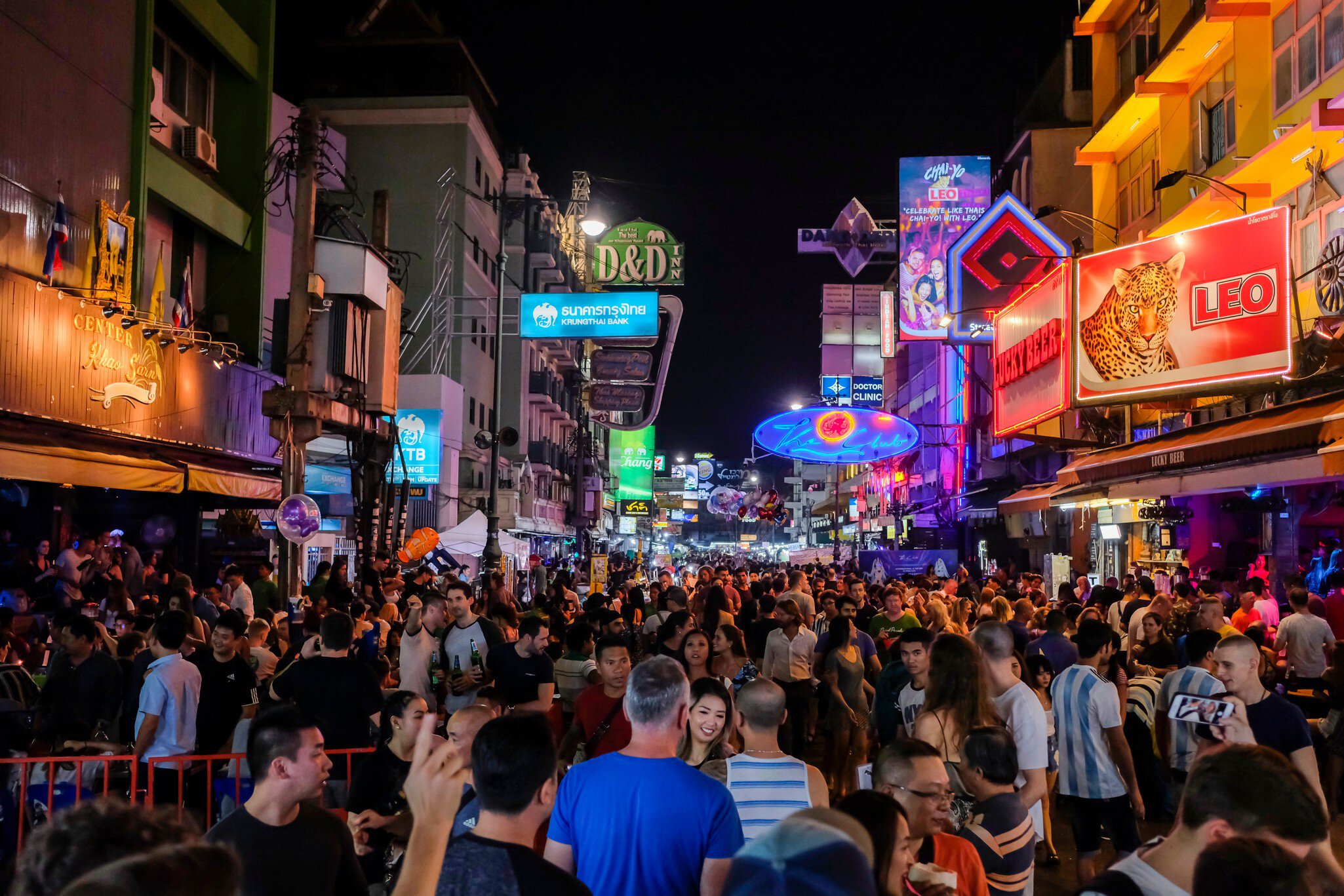 Hundreds of people walk at night along a road that has buildings with neon signs and billboards overhead.