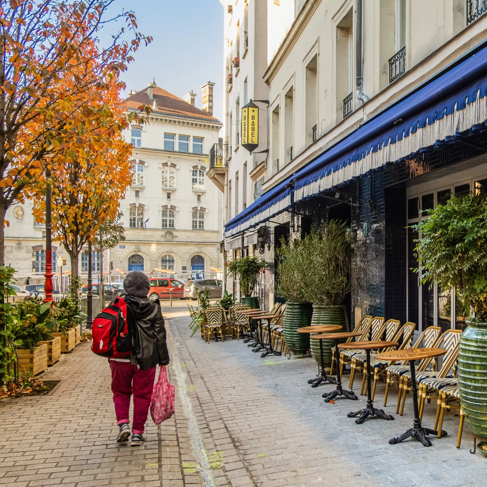 A person walks past the blue awning and outdoor tables of a hotel on the right side and orange-foliage trees on the left side