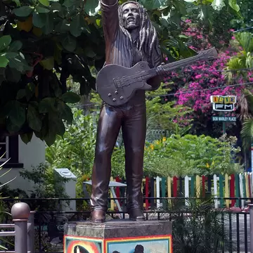 Bronze figure of a musician with dreadlocks raising one arm and holding a guitar, on a painted plinth under trees.