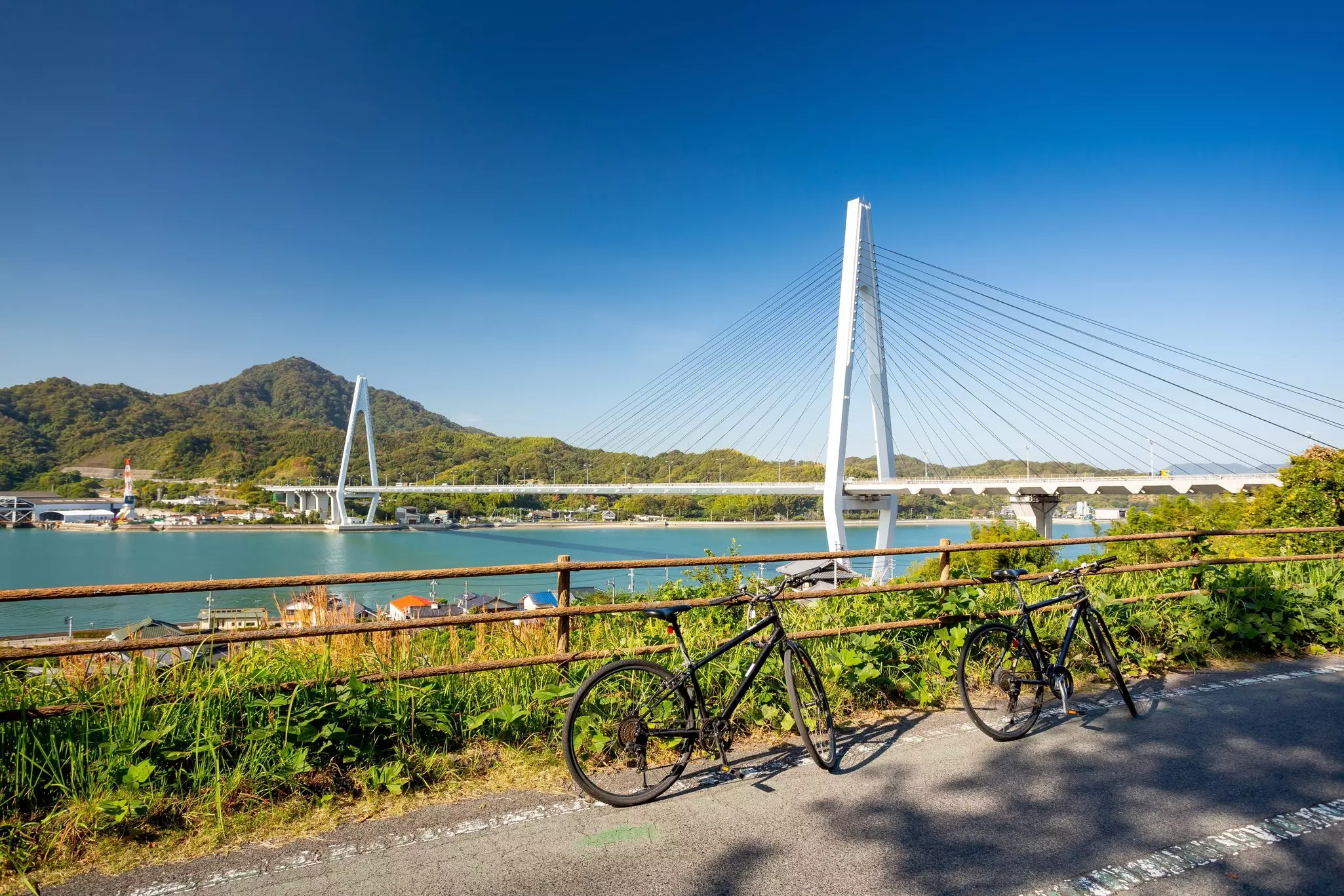 Two bicycles lean against a railing on a path above a waterway crossed by a large suspension bridge in Japan.