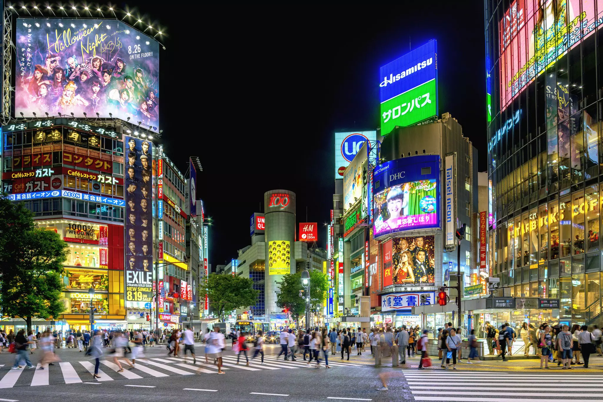 People crossing a large crossroads at night with neon signs lit up on the buildings all around.