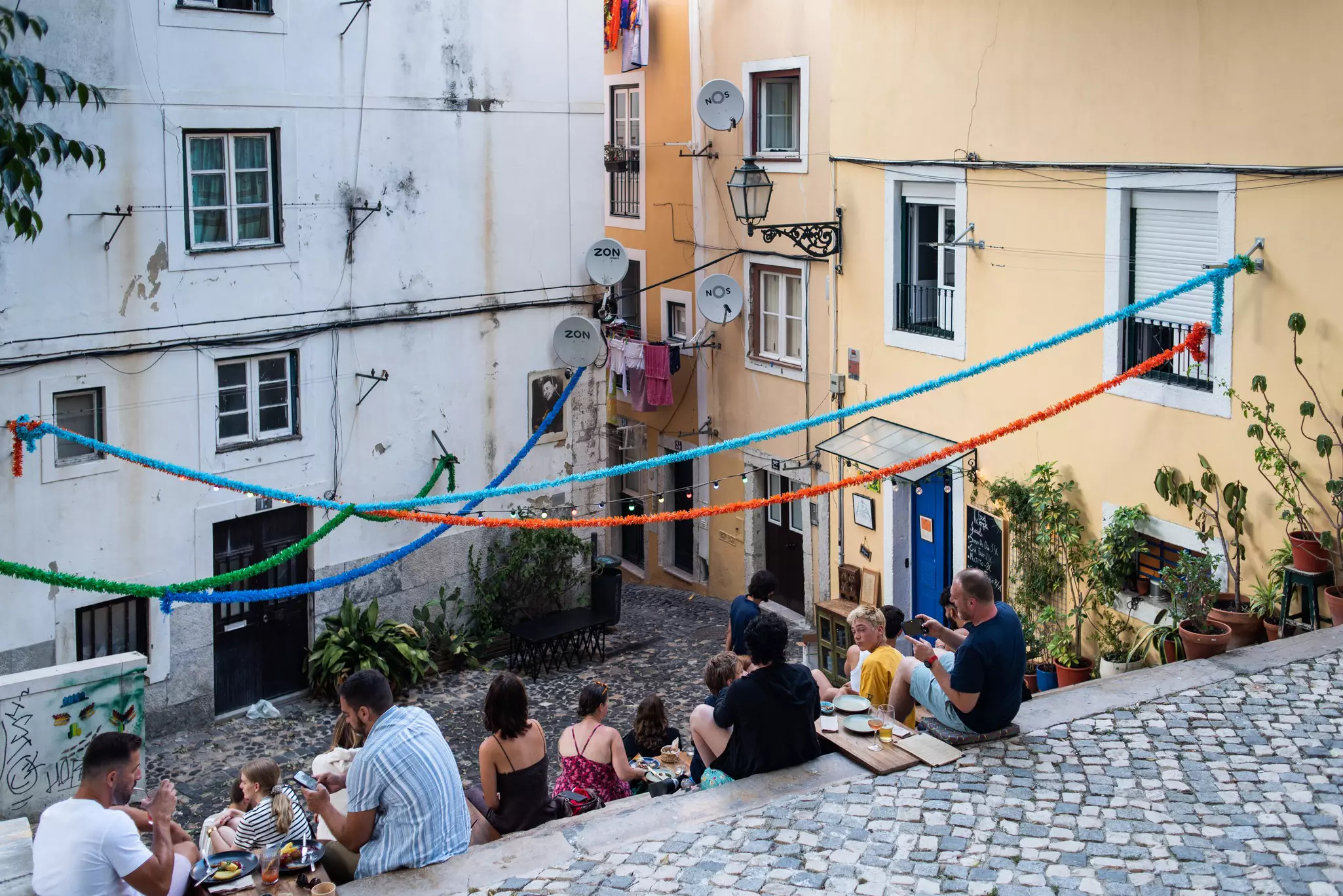 People are seen from behind sitting on steps in a city neighborhood. Many are eating.