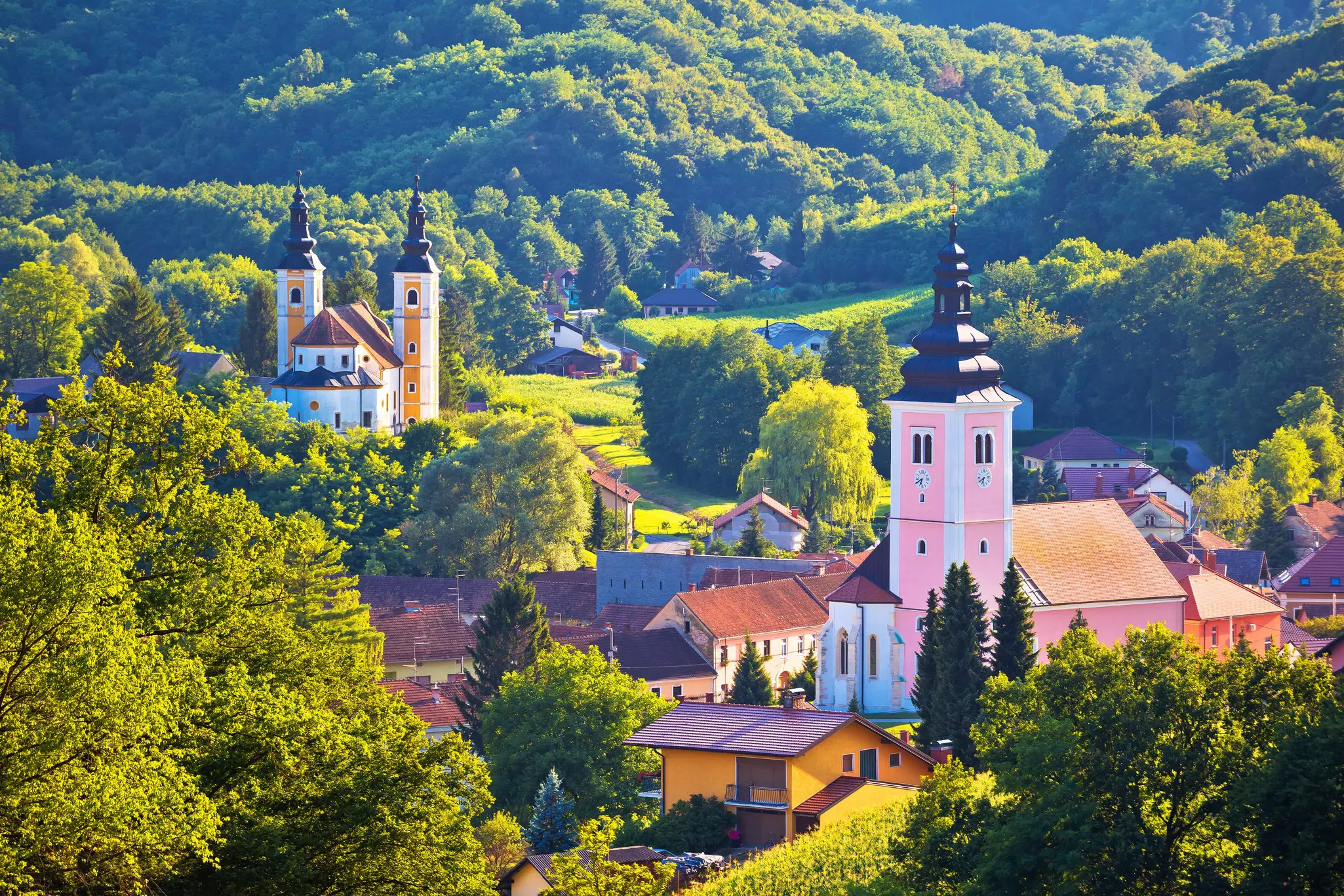 A village in a green valley in warm light with strong shadows; a building with a pink tower is in the foreground and a building with two yellow towers is in the background.
