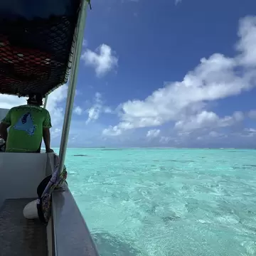 Clear turquoise shallows running out to a distant reef line, seen past the skipper of a small awning-covered boat.