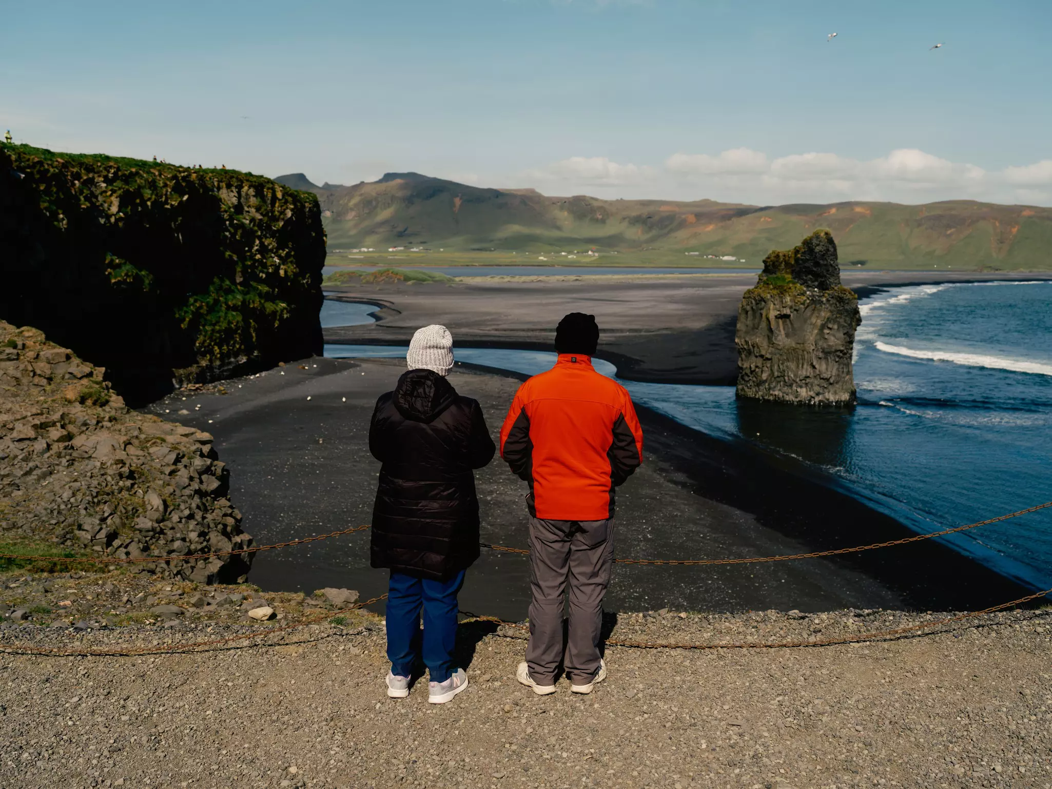 People in heavy coats stand on a cliff above a black sand beach; there is a large rock off the shore.