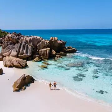 Aerial view of a small couple on white sand beside huge granite boulders and clear turquoise shallows on La Digue