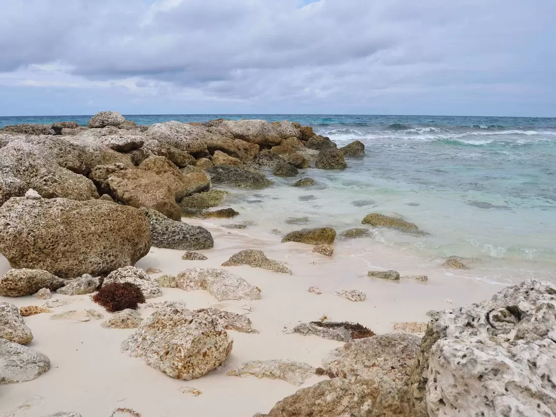 Pitted coral rocks on white sand at the edge of turquoise water under grey cloud, Blue Lagoon Island, Bahamas