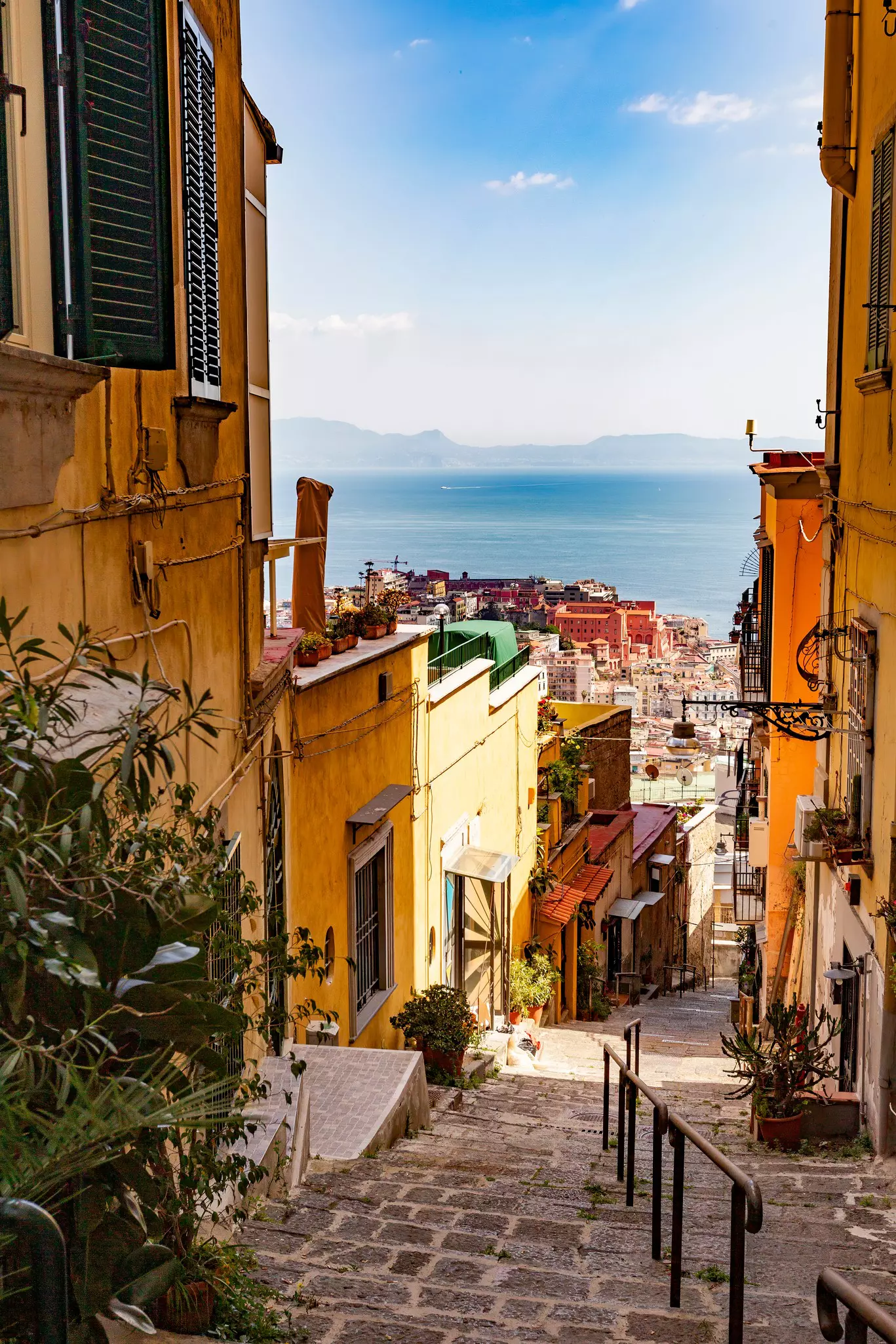 A cobbled street leading downhill between residences to a tightly packed seaside city below.