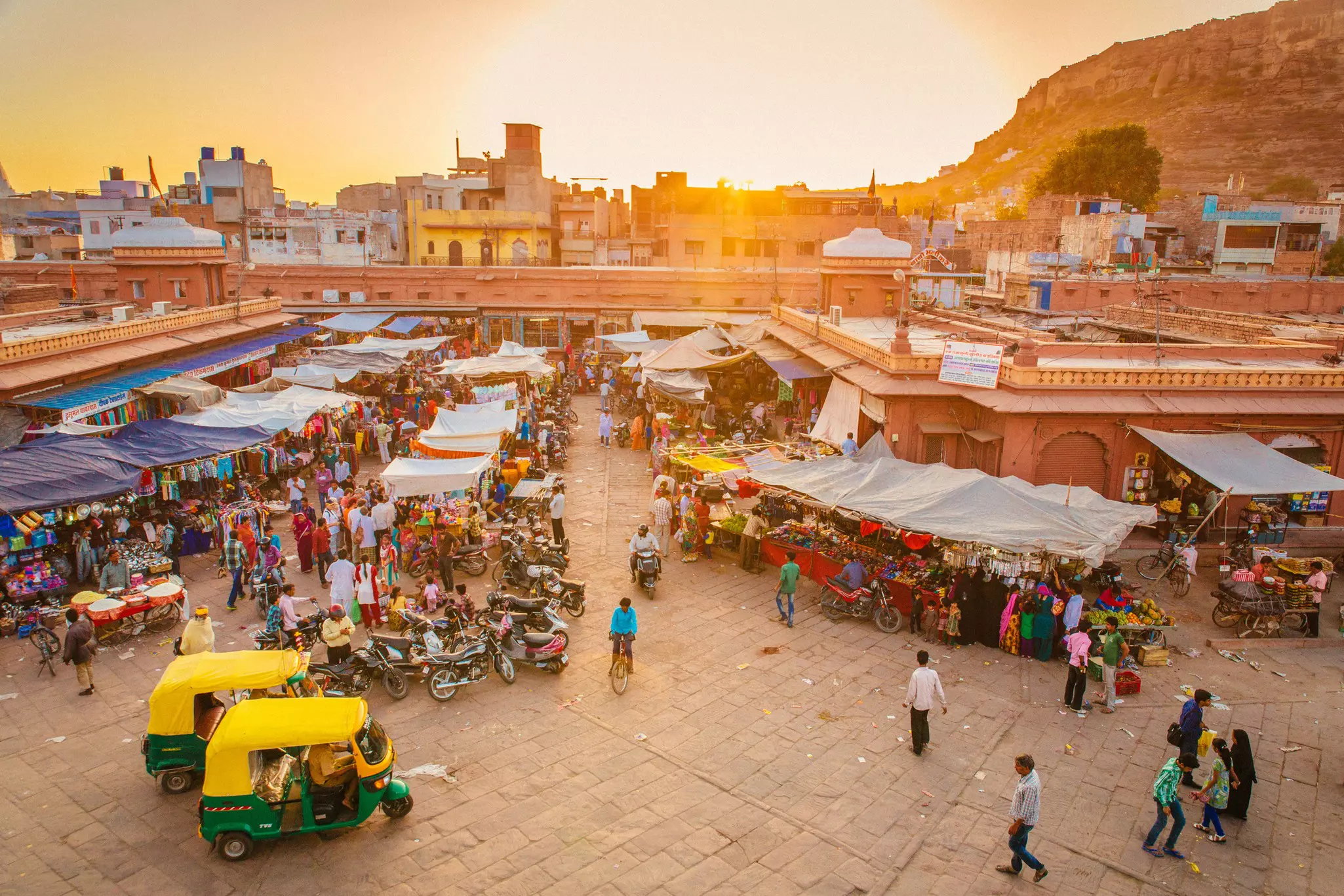 Rickshaws and motorcycles wait near stalls at a busy market in a city with red walls