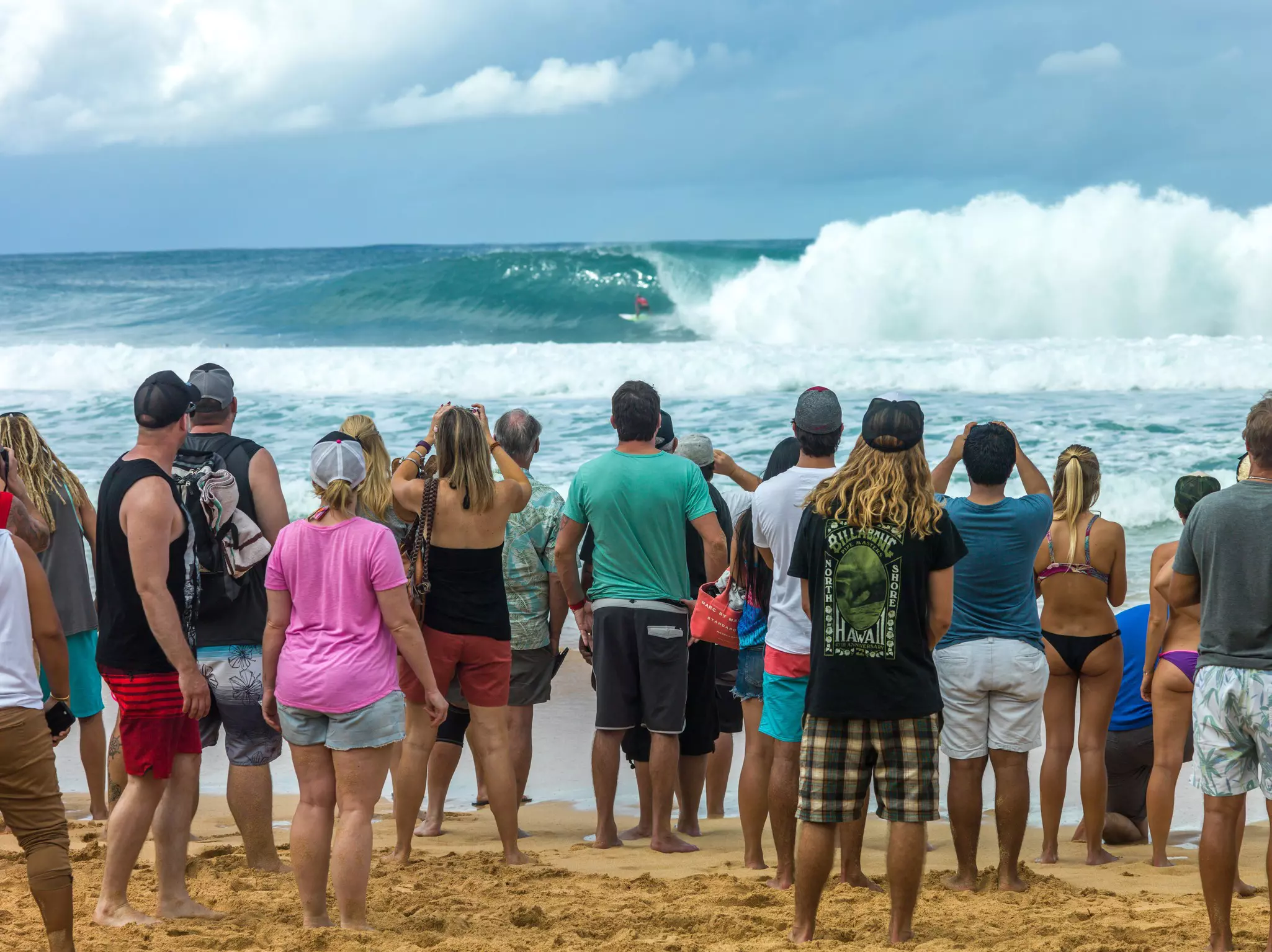 People stand on beach watching surfers on a huge wave offshore.
