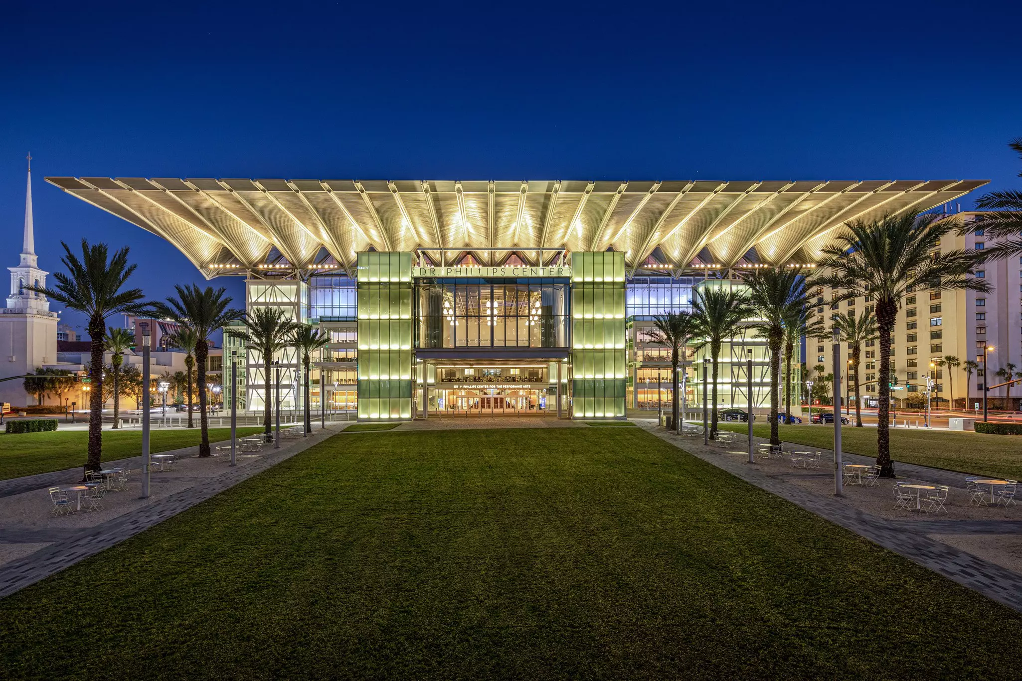 Exterior of glass-fronted building reading "Dr Phillips Center" above the entry with green grass and palm trees lining the front entry. The building is lit up at nighttime.