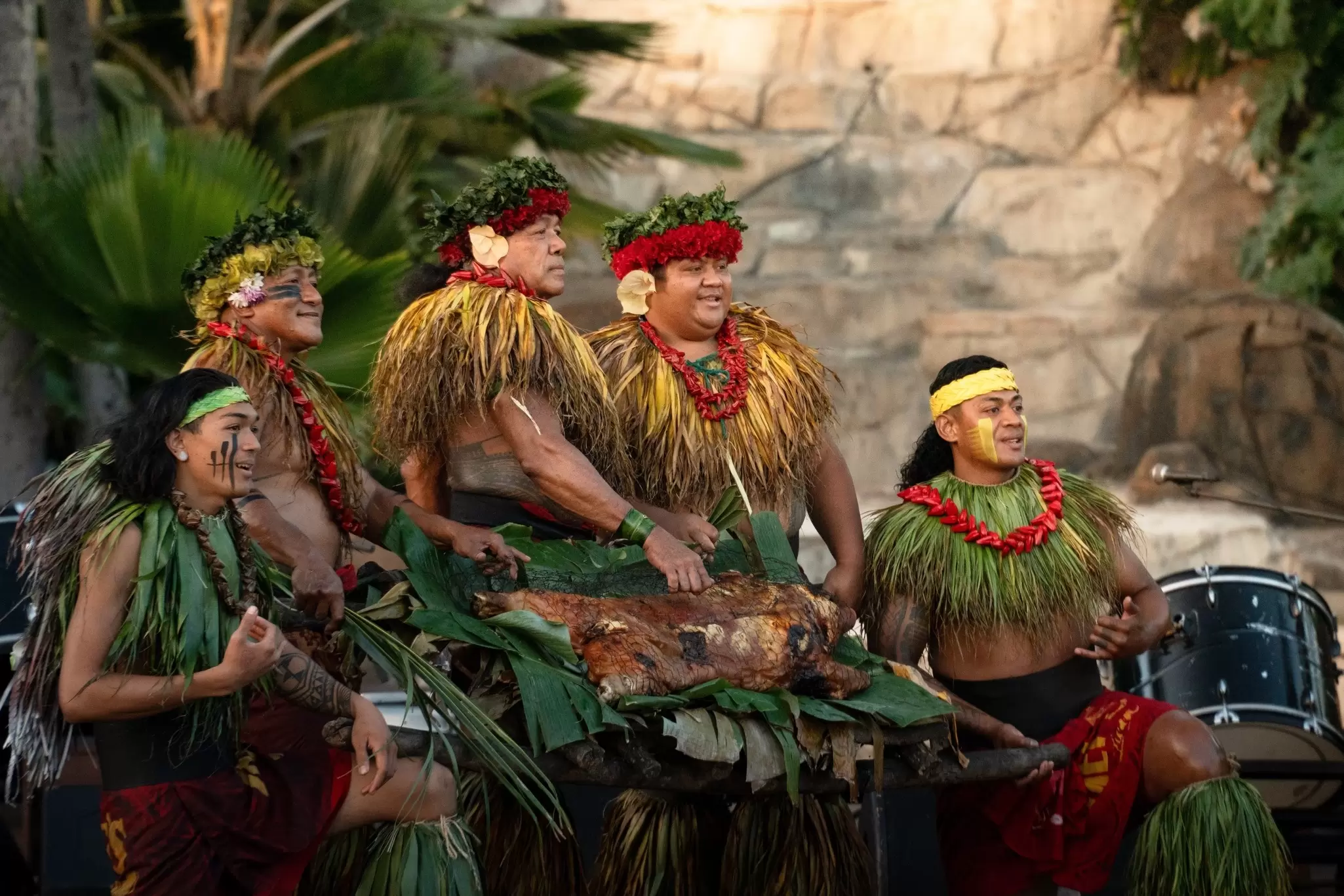 Five men dressed in traditional Hawaiian clothing hold a cooked pig