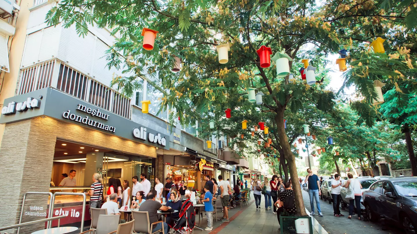 A tree-lined street with many people passing by. Some are sat outside a cafe to the left. The tree has colourful lanterns hanging from the branches