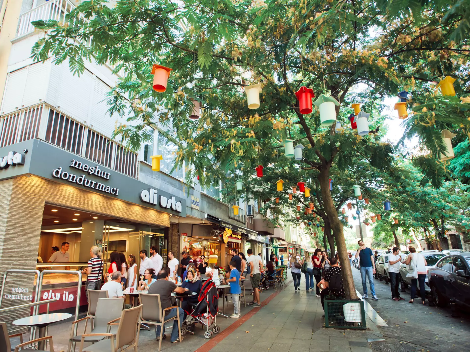 A tree-lined street with many people passing by. Some are sat outside a cafe to the left. The tree has colourful lanterns hanging from the branches