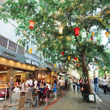 A tree-lined street with many people passing by. Some are sat outside a cafe to the left. The tree has colourful lanterns hanging from the branches