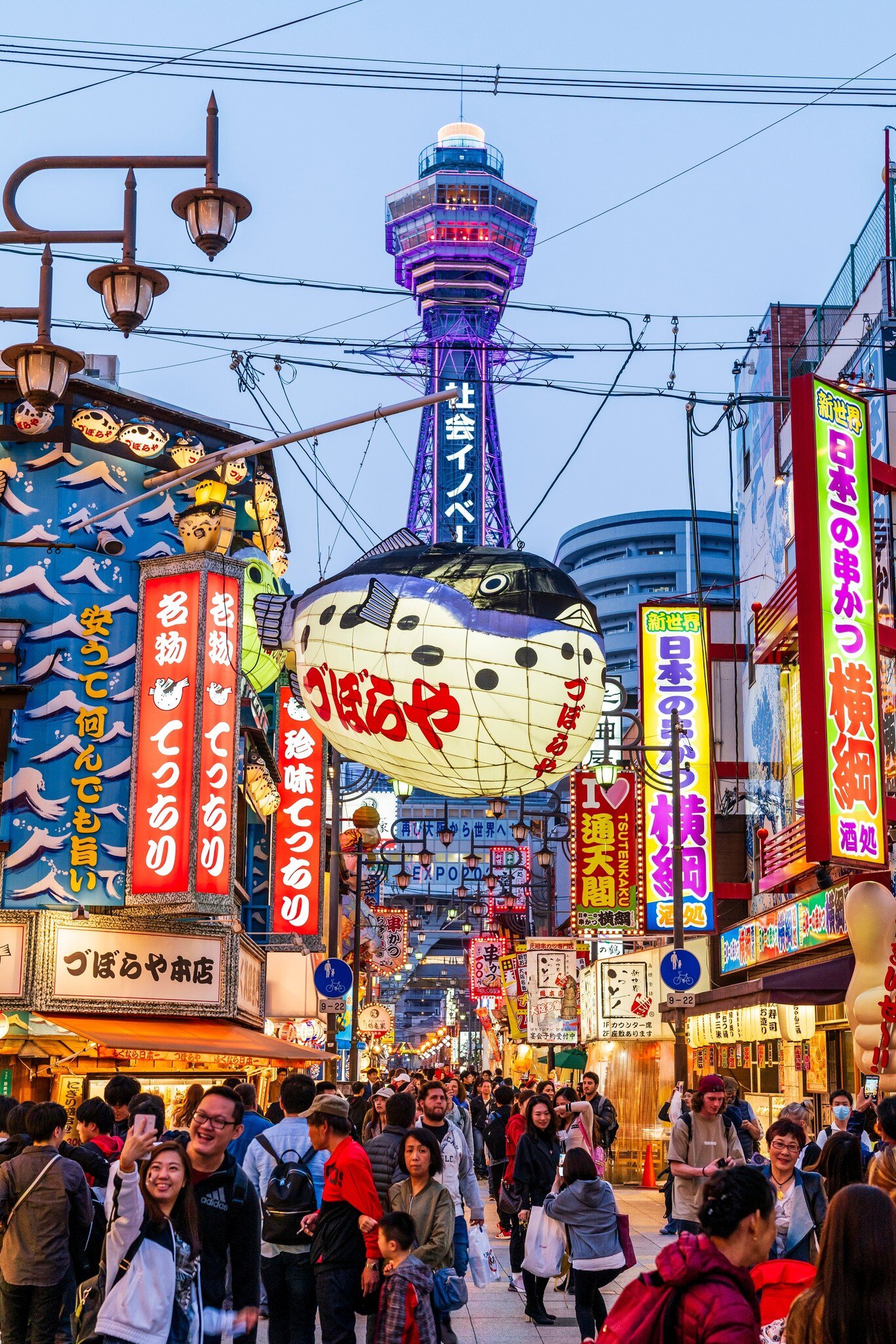 Shinsekai, Osaka, Japan. April 6 2019. Street view, crowded pedestrian precinct with the Tsutenkaku Tower towering over Shinsekai at blue hour. Illuminated Puffer fish lantern, Zuboraya. restaurant, License Type: media, Download Time: 2026-05-28T19:31:03.000Z, User: LP_NZhan, Editorial: true, purchase_order: 65050 - Digital Destinations and Articles, job: App Content, client: Lonely Planet App, other: Nadine Zhan
