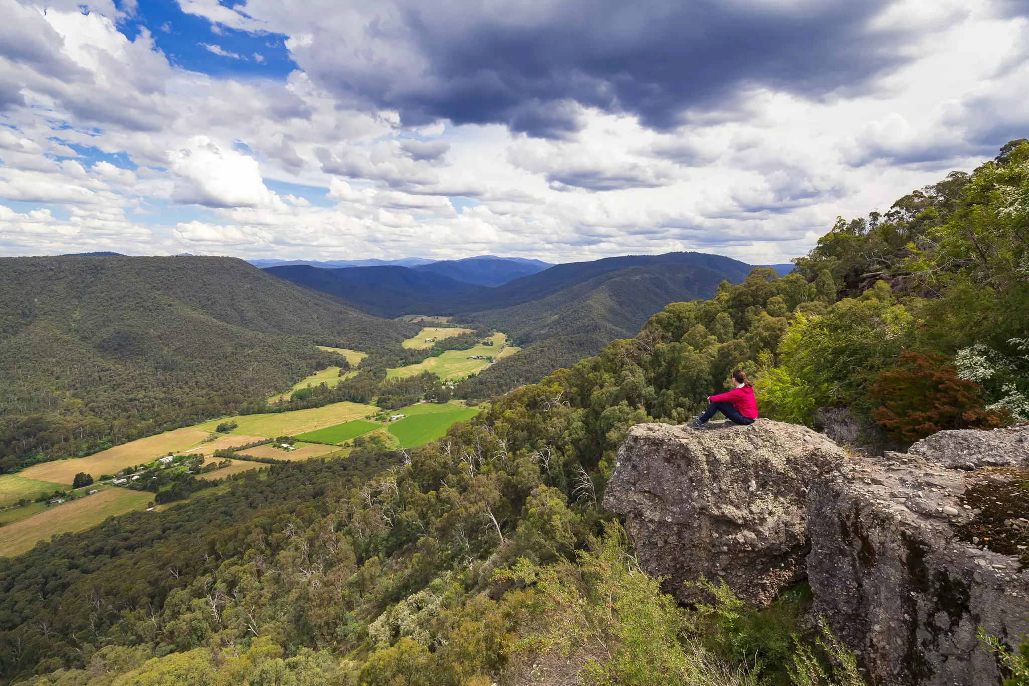 A woman sits on a rocky outcrop on a mountain looking out at a view of valleys and fields below