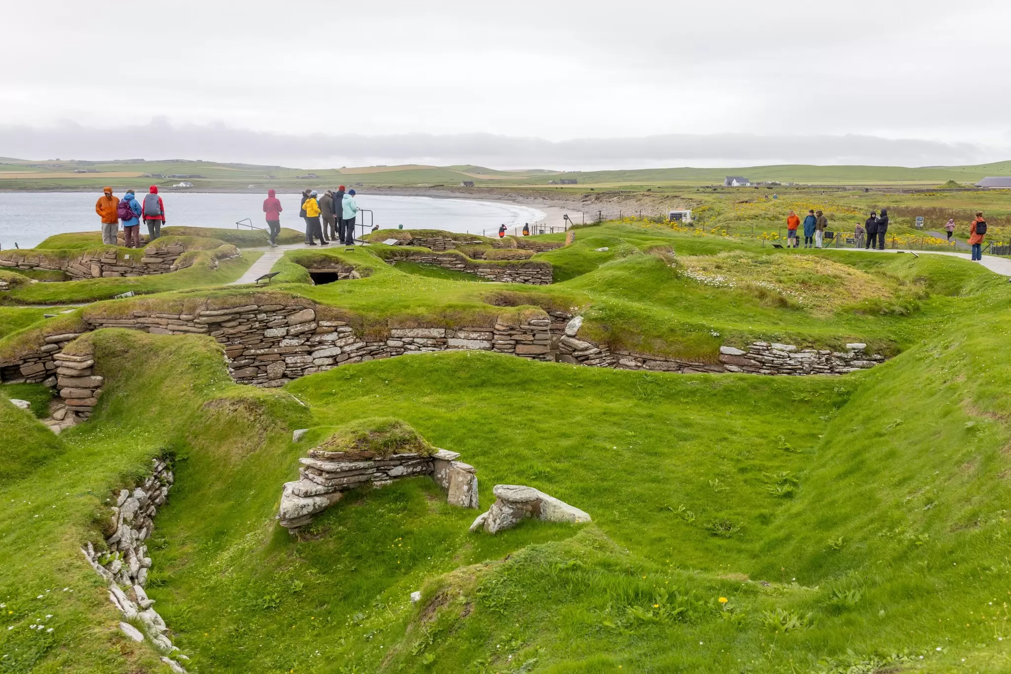A series of stone houses built into green mounds of earth on the edge of an island with the grey sea churning nearby