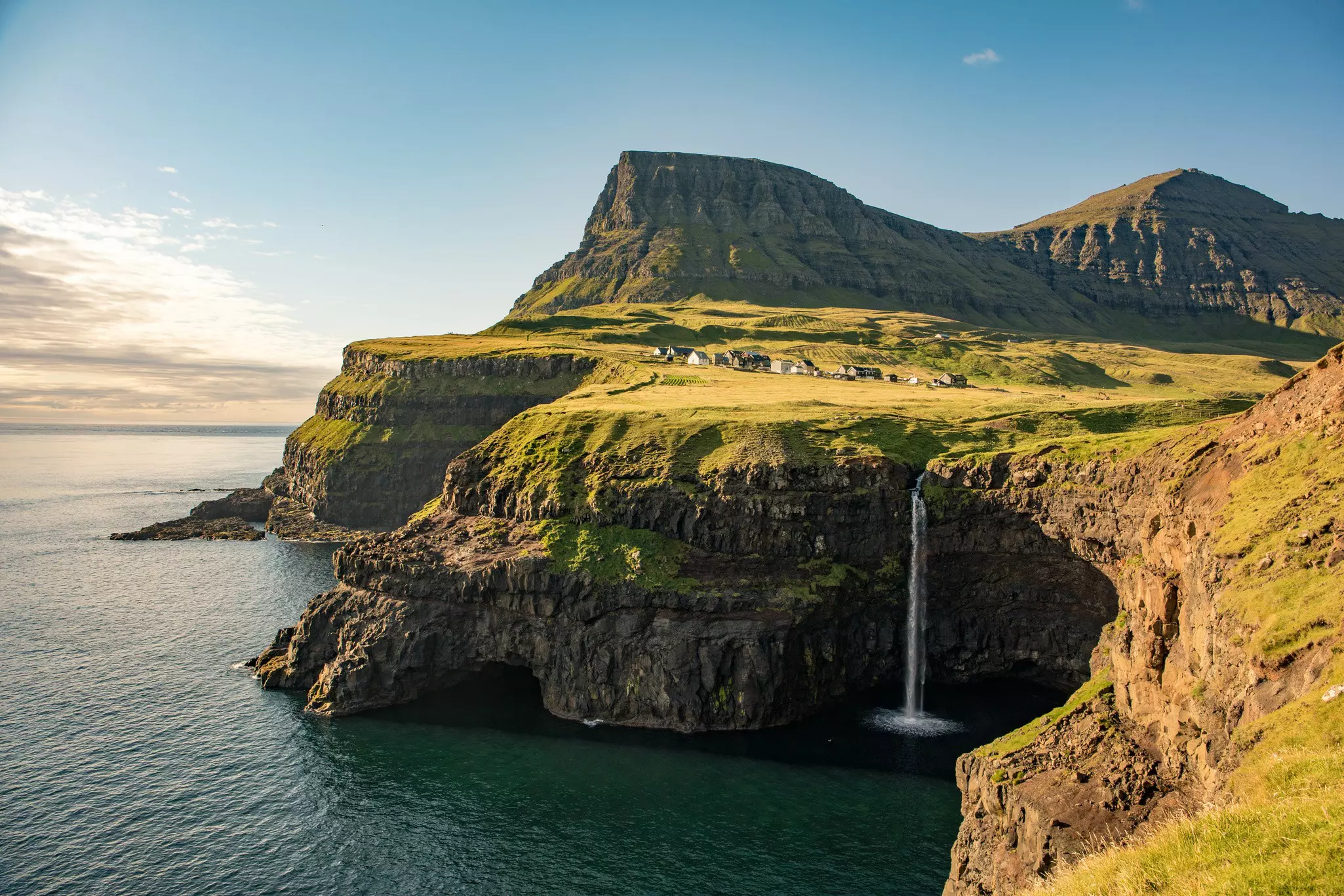 A waterfall cascades down off a green cliff top and over a cave mouth into the sea.