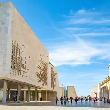 A large limestone building raised on slender columns, its facade deeply carved into an irregular relief, with people crossing the paved square in front.