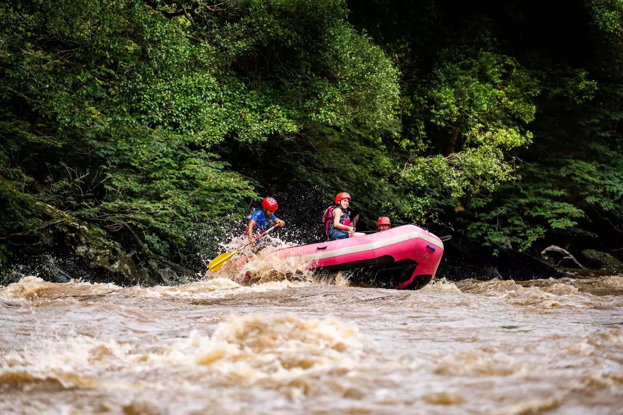 Rafters with paddles in a small red boat travel down a choppy stretch of river surrounded by thick foliage.