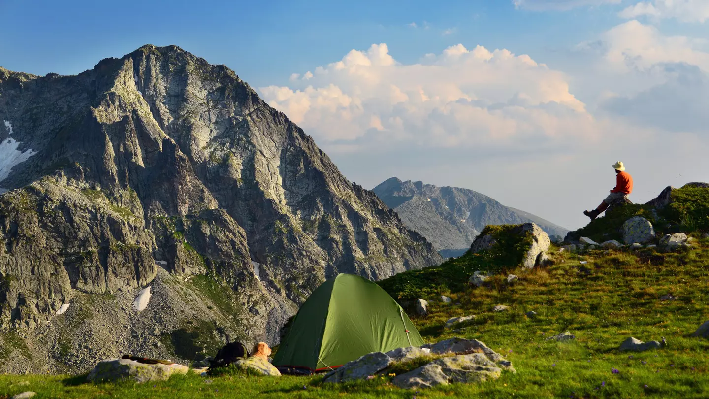 A man sits on a rock and looks at a mountain vista. A green tent is seen in the foreground.