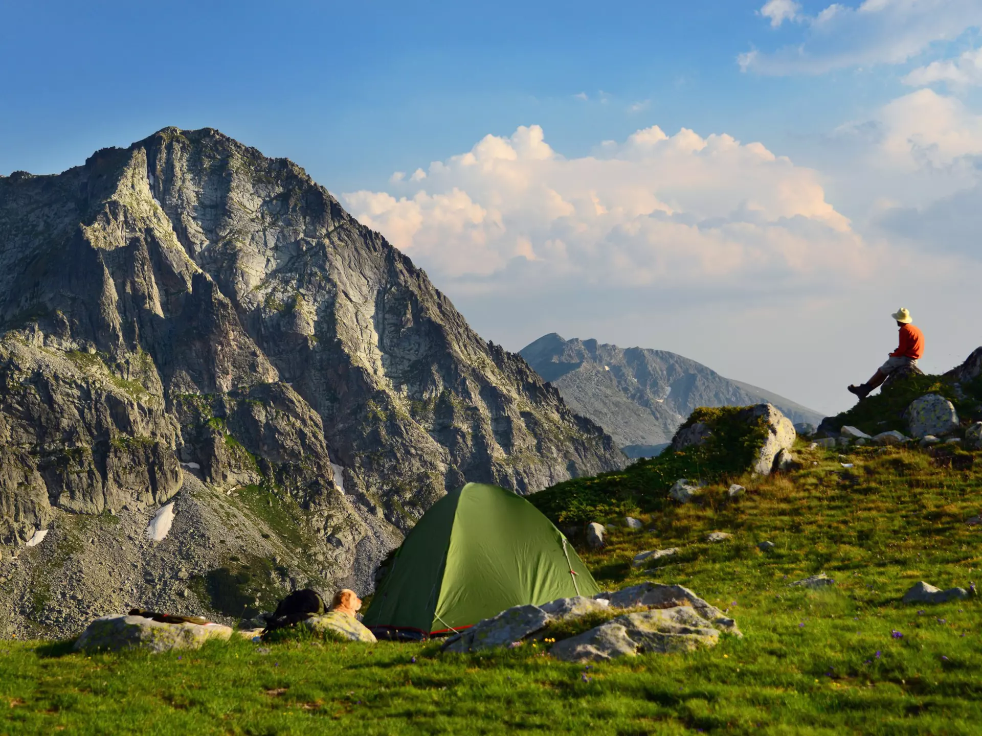 A man sits on a rock and looks at a mountain vista. A green tent is seen in the foreground.