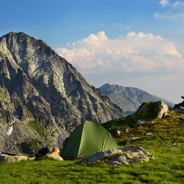 A man sits on a rock and looks at a mountain vista. A green tent is seen in the foreground.