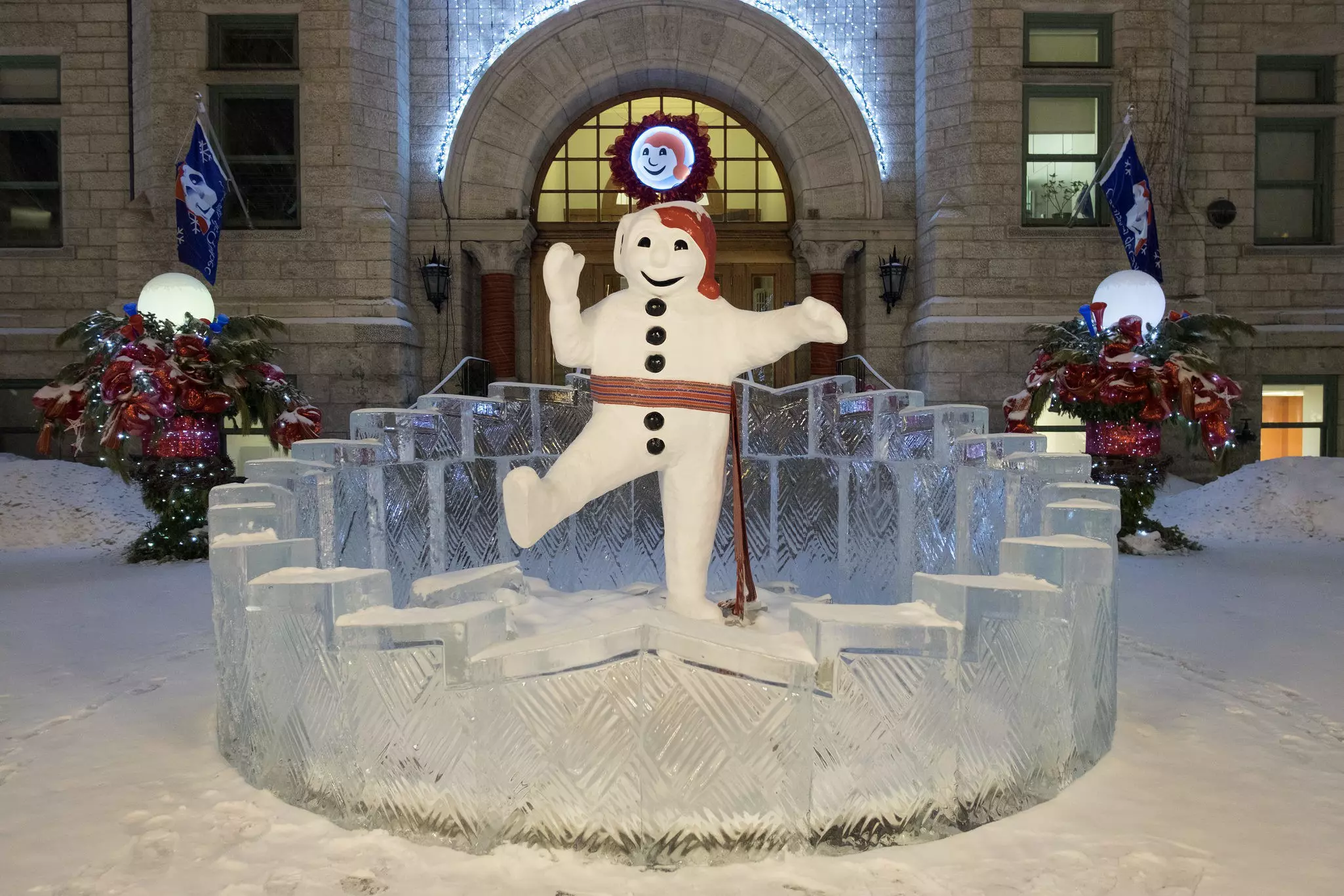 Statue of a jaunty, humanoid snowman standing on an ice sculpture surrounded by snow and kicking one leg up at nighttime.