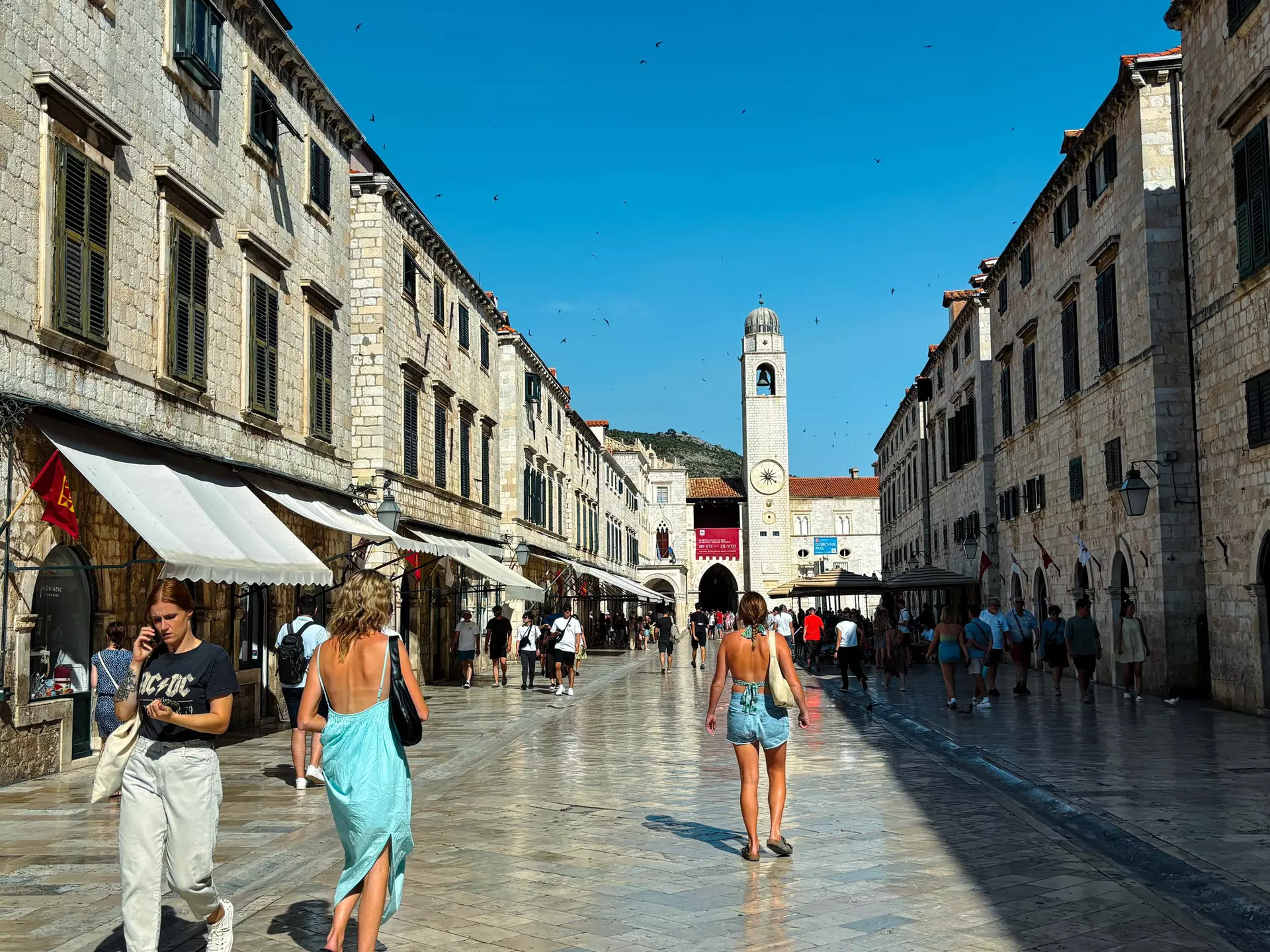 People walk down a pedestrianized street with a tall bell tower in the distance in Dubrovnik, Croatia.