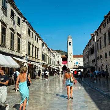 People walk down a pedestrianized street with a tall bell tower in the distance in Dubrovnik, Croatia.