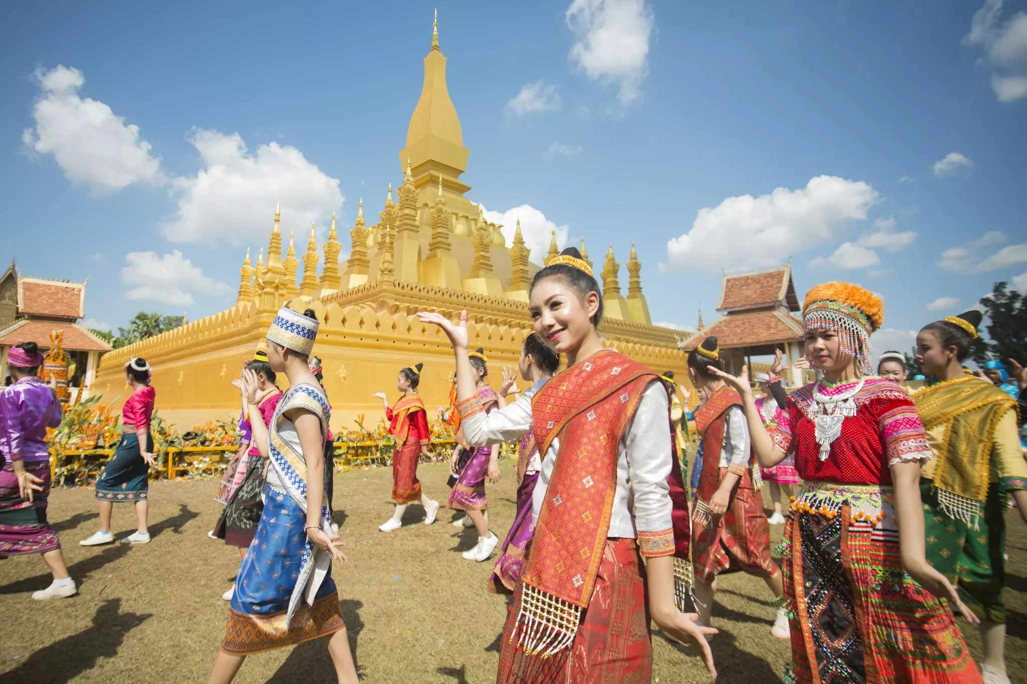People in traditional costumes make hand gestures as they participate in a parade past a temple in a city street.